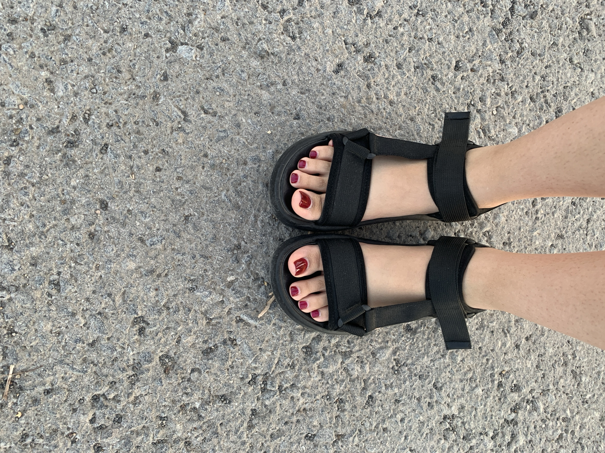 A pair of feet, adorned with dark red nail polish, are visible from a top-down perspective. The feet are wearing black platform sandals with adjustable straps. They are positioned on a rough, grey concrete surface that appears to be a sidewalk or road in the city of Asha, Russia. The concrete shows texture and small embedded pebbles. The lighting suggests it is daytime.