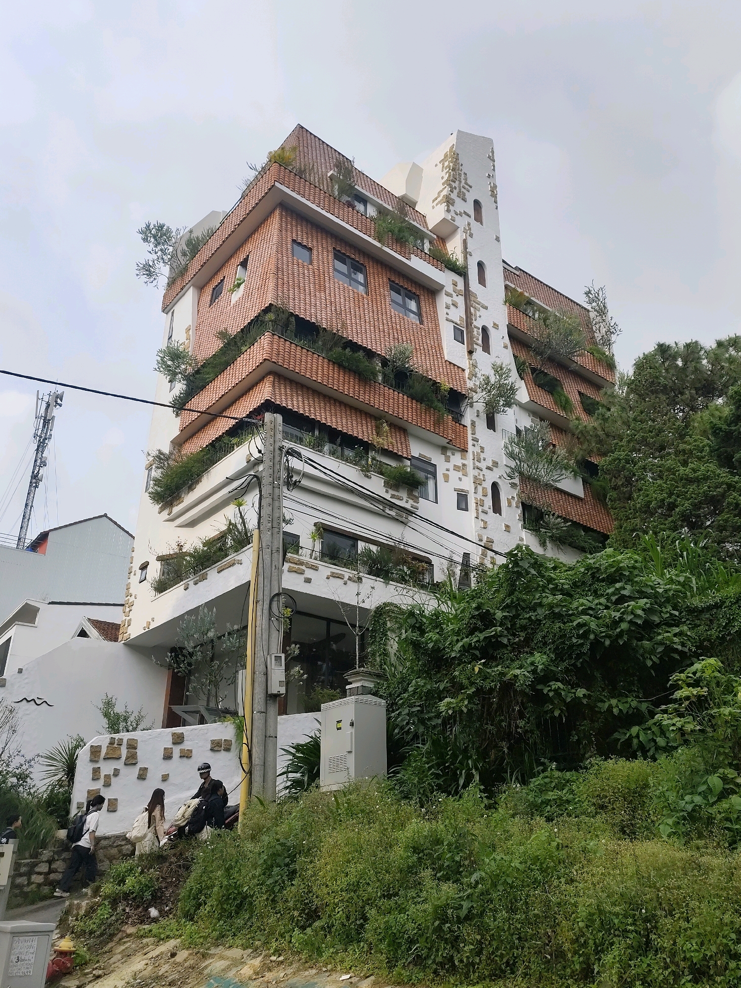 In Lâm Viên - Đà Lạt, Vietnam, a multi-story building with a unique architectural design is pictured. The building features a stepped facade covered in terracotta tiles, with several balconies adorned with lush greenery and small trees. A prominent white tower with stone accents, resembling a castle turret, rises from one side of the building. The scene also includes a cell tower in the distance, overhead electrical wires, and a dense green hillside covered in various plants and trees. At the bottom of the image, on a stone pathway, three individuals are visible. One person, wearing a helmet and sitting on a motorcycle, interacts with two others who are walking up the steps. One of the walkers carries a backpack, and the other has a tote bag slung over their shoulder. The overall atmosphere is one of a slightly overcast day in a verdant urban environment.