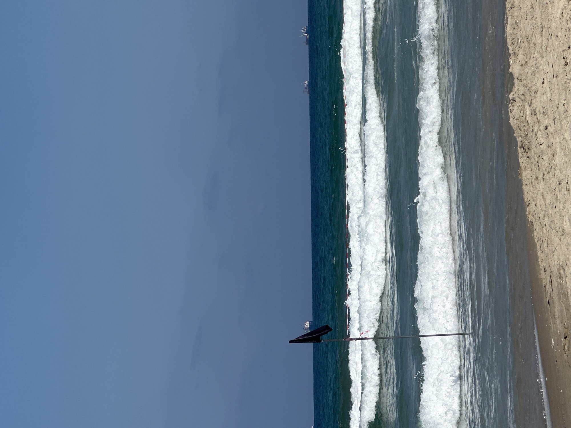 A wide shot captures a serene beach scene in Kiryat Yam, Israel, under a clear blue sky. The foreground features a sandy beach with subtle footprints. Moving towards the background, the vibrant blue sea is marked by three distinct lines of white-crested waves rolling towards the shore. A black flag stands planted in the shallow water near the beach, attached to a thin pole. In the distance, several ships are visible on the horizon, dotting the vast expanse of the ocean.