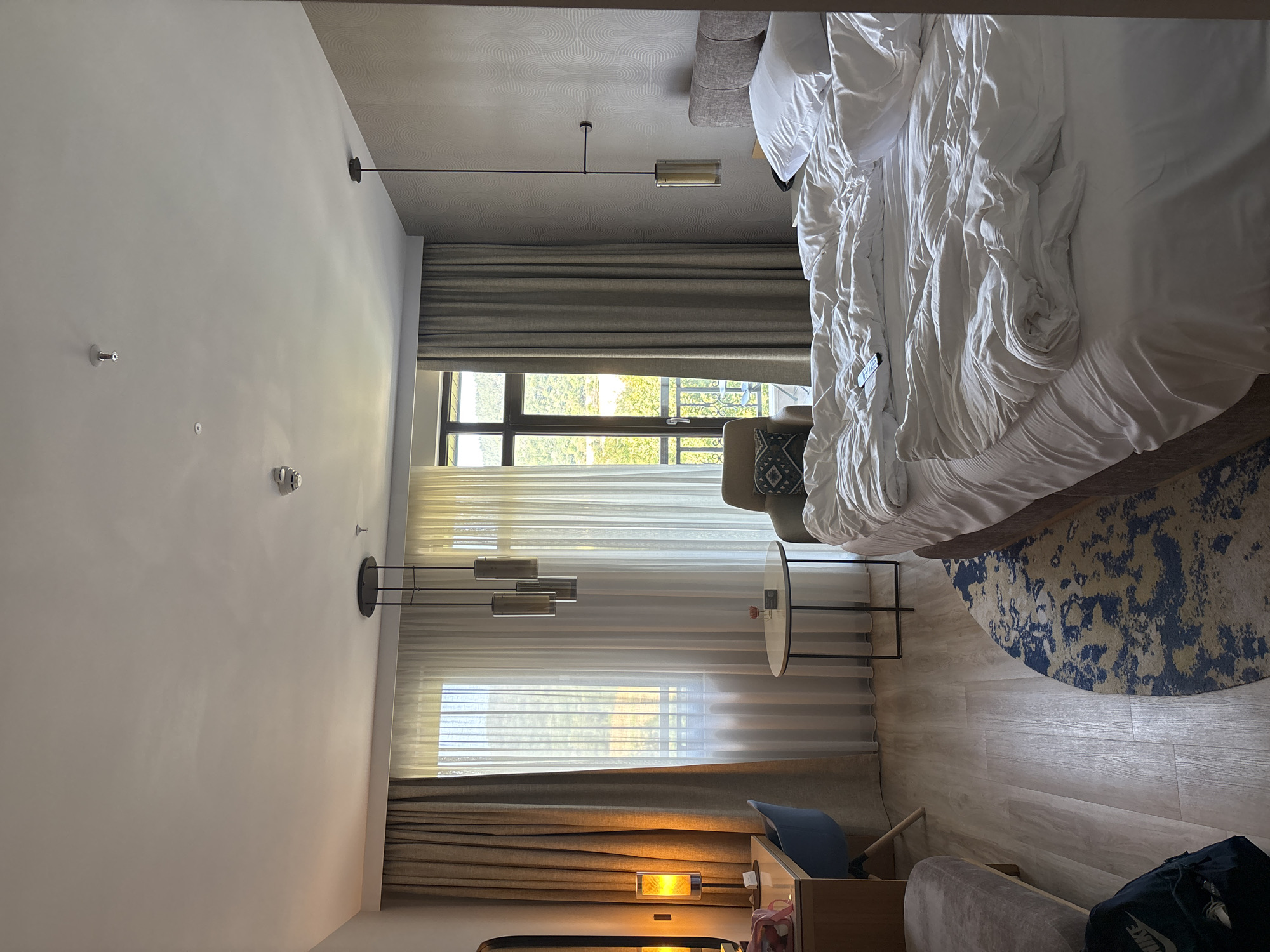 The image depicts a hotel room in Razliv, Bulgaria. The room contains a large, unmade bed with white linens and a crumpled duvet. To the left of the bed is a small, round side table with a vase containing a single pink flower. Further left, a desk area is visible with a wooden desk, a blue chair, and a lamp emitting a warm yellow light. Behind the desk, a tall window is covered by two layers of curtains: thick brown curtains on the outside and sheer white curtains behind them, allowing natural light to filter through. Through the window, a balcony railing and lush green trees can be seen. Above the bed, a modern pendant light fixture hangs from the ceiling. To the right of the bed, a patterned wallpaper covers the wall, and a modern wall lamp is mounted above a headboard. A patterned rug with blue and white designs is on the wooden floor between the bed and the desk area. A black backpack with a Nike logo is on the floor in the lower left corner. The overall impression is that of a comfortable, well-lit hotel room.