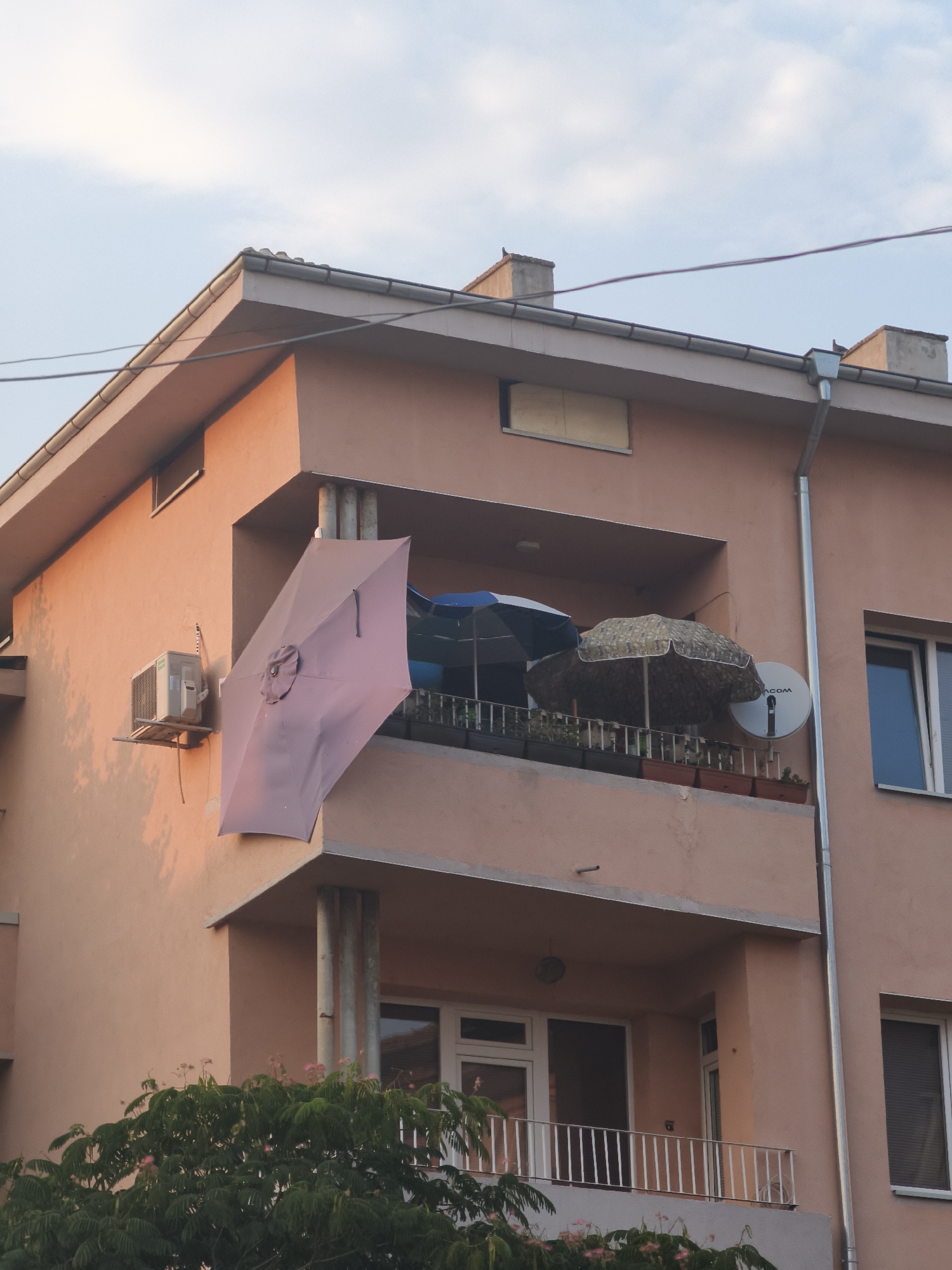 The image shows a building with a balcony in Lovech, Bulgaria. The building is painted in a peach color. On the balcony, there are several umbrellas, some of which are open. There is also a satellite dish and some potted plants. An air conditioning unit is mounted on the wall of the building. The sky is visible in the background with some clouds. There is also some foliage from a tree in the foreground. The scene appears to be a residential building.