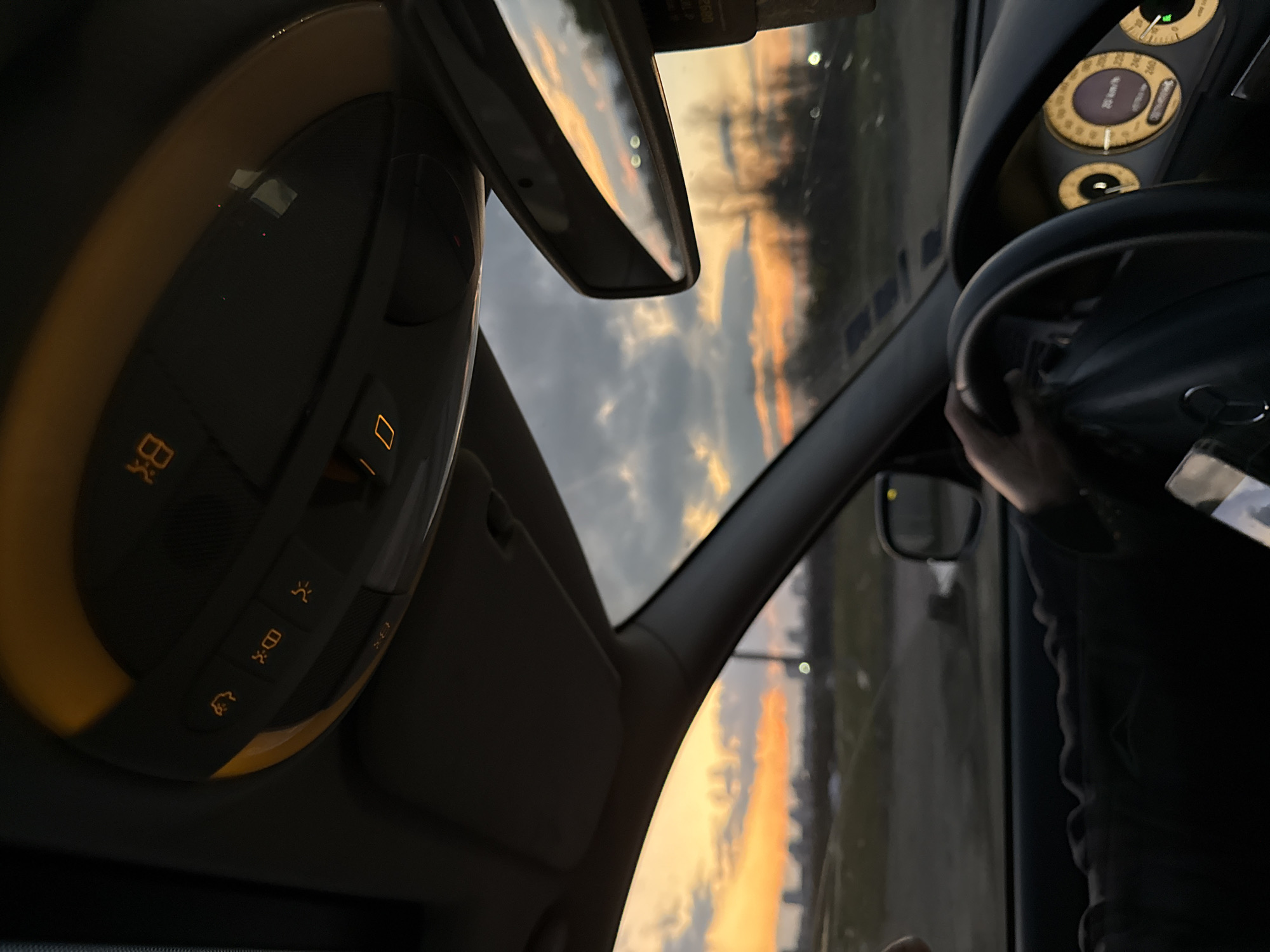 The view from inside a car shows a sunset reflected in the rearview mirror and windshield. The car's interior features a control panel with illuminated icons and a steering wheel being held by a person's hand. The speedometer reads 26 km/h. Outside, the background shows an urban landscape with buildings silhouetted against the sky. The location is likely Sofia, Bulgaria, given the urban landscape and European-style dashboard.