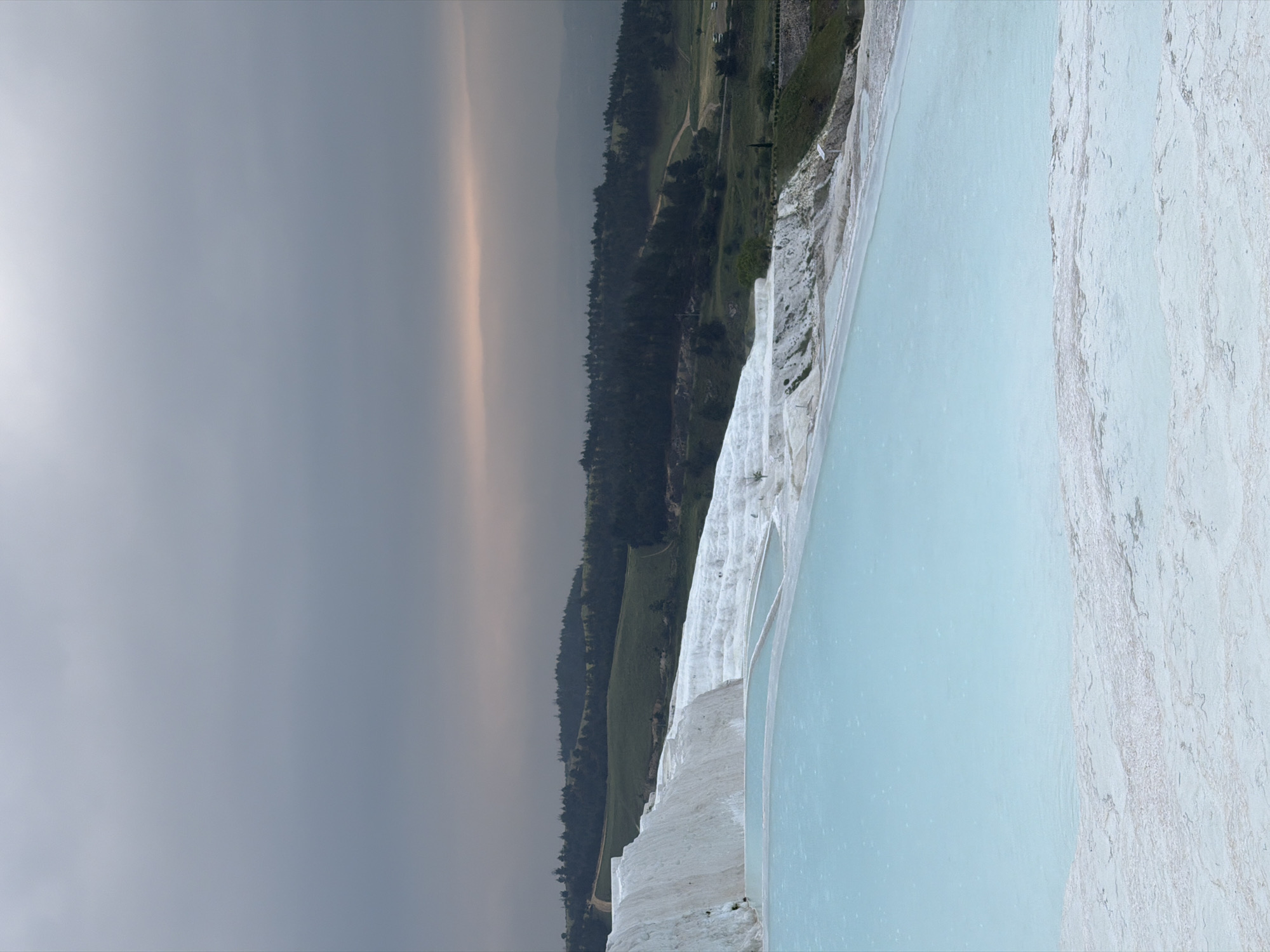 The image captures Pamukkale's thermal pools in Turkey. The pools are visible in the right portion of the frame, where water is light blue and the terraces are white. Adjacent to the pools, a strip of green vegetation and a tree line are also present. The sky, occupying the left side of the image, is gray and overcast.