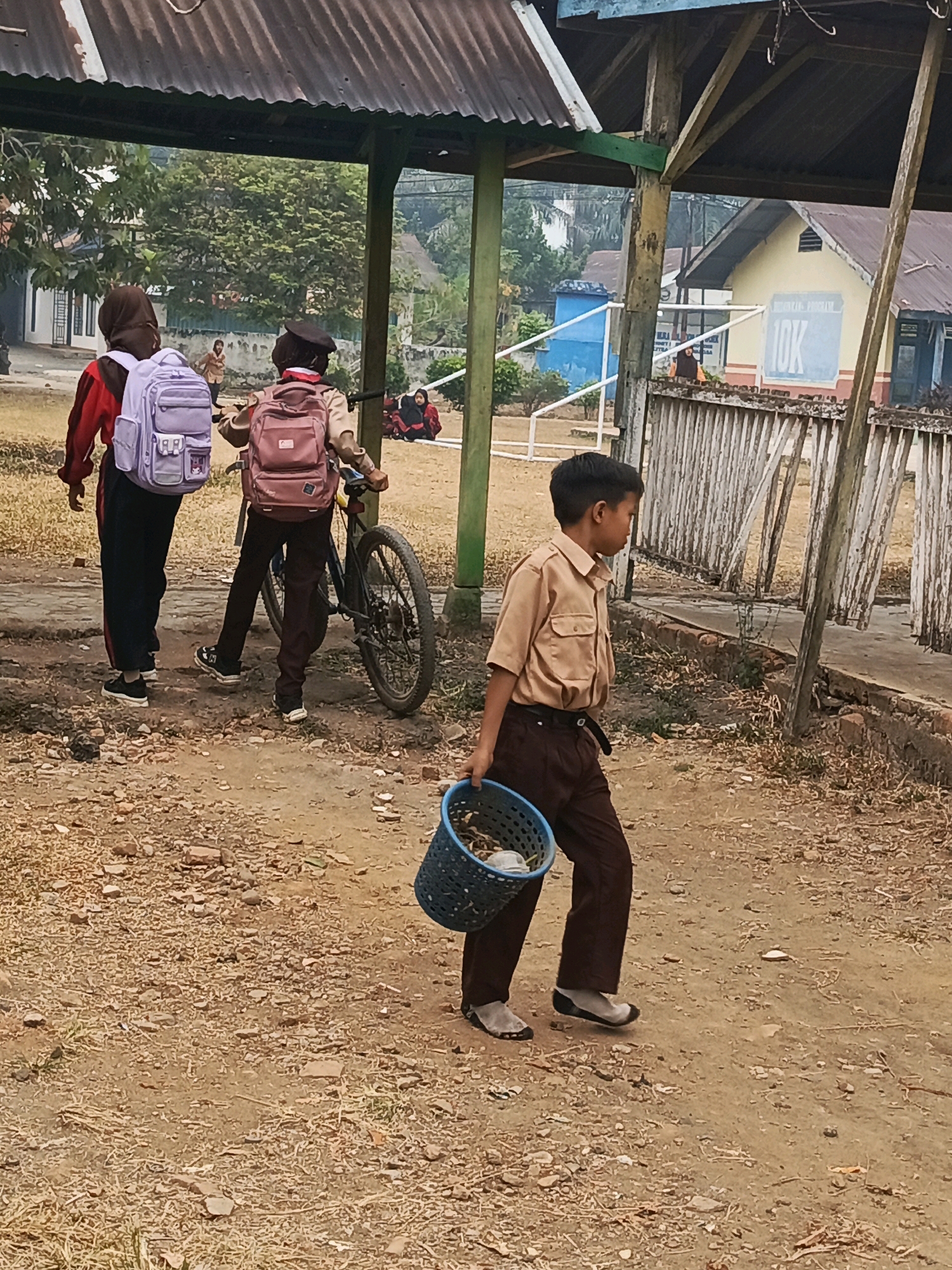 The image depicts an outdoor scene likely on the grounds of a school in Megang Sakti II, Indonesia. In the foreground, a young boy wearing a light brown collared shirt and dark pants walks towards the right, carrying a blue plastic basket filled with what appears to be dried leaves and other debris. He is wearing grey socks and dark, slip-on shoes. Behind him, two students are walking to the left. The student closer to the viewer wears a light brown uniform and a backpack, pushing a bicycle. The student further left wears a red jacket, dark pants, and a light purple backpack. They are walking on a dirt path with sparse dry grass and scattered stones. In the background, several school buildings with a light yellow facade are visible, along with a blue structure. Wooden fences and trees are also present, suggesting a rural or semi-urban school environment. The overall lighting indicates it is daytime, possibly late afternoon, with soft shadows.