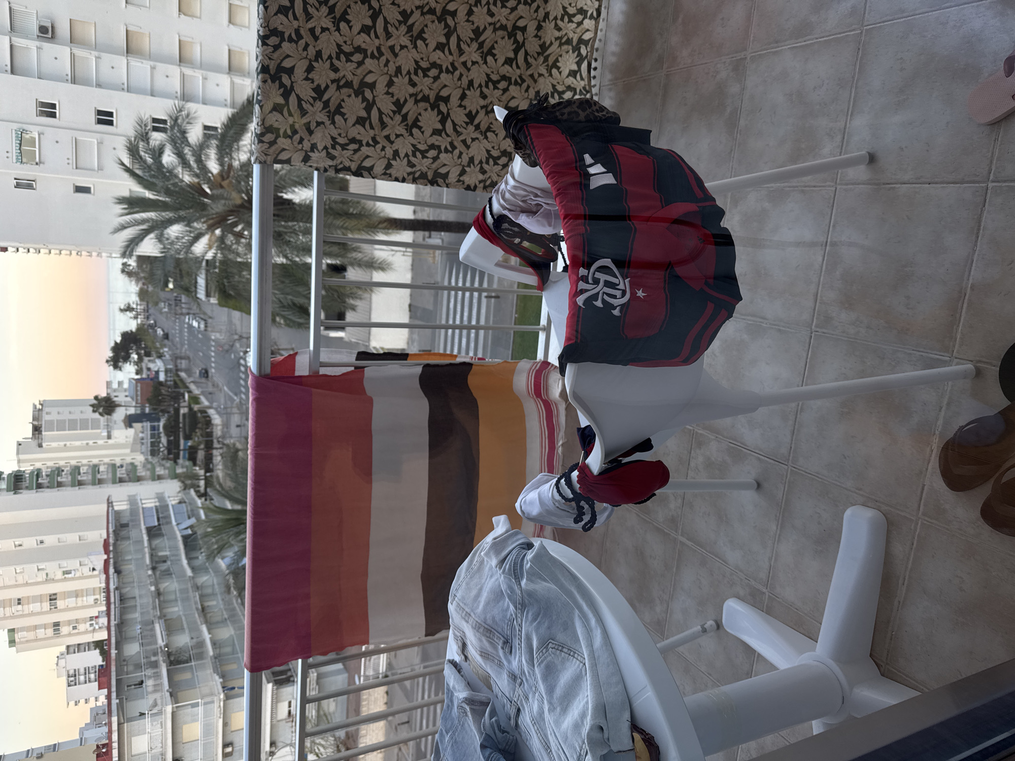 The image, taken in Grau I Platja, Spain, depicts a balcony scene with a clothes drying rack holding a variety of items. A striped towel hangs on the left side of the rack, while a blue denim jacket rests on the bottom. On the right, a red and black Flamengo soccer jersey is draped over the rack, along with other miscellaneous clothing items.
The balcony offers a view of the city, including buildings, a street, and palm trees. The balcony flooring is tiled. Two pairs of footwear are visible near the edge of the balcony: a pair of sandals and a pair of brown shoes. A curtain with a leafy pattern hangs partially behind the drying rack. The image is taken from a slightly elevated position, looking down onto the scene.