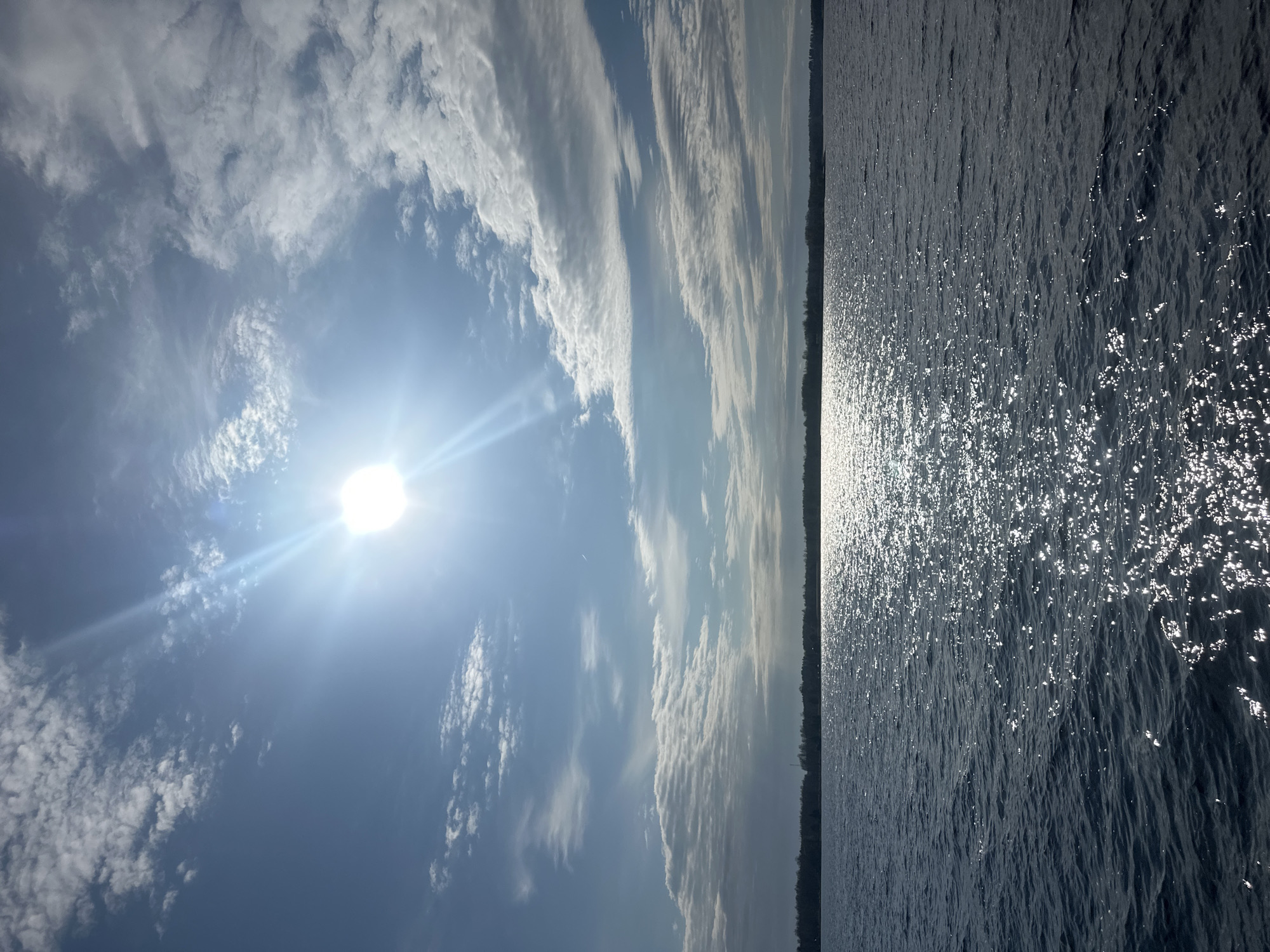 The image captures a bright, expansive outdoor scene, divided horizontally between sky and water by a distant treeline. The upper two-thirds of the frame display a vibrant blue sky dotted with scattered clouds, ranging from wispy formations to more textured patterns. The sun, a brilliant disc, is positioned on the left side, emitting strong rays and a prominent lens flare that illuminates the sky.
A narrow, dark band of land, likely a distant forest or shoreline, forms the horizon, stretching across the entire width of the image. Below this horizon, the lower third of the photograph reveals a large body of water, its surface disturbed by numerous small ripples. The intense sunlight reflects brilliantly off the water, creating a sparkling, shimmering effect with numerous white highlights and streaks across its dark surface.
The scene depicts a sunny day, likely midday or early afternoon given the sun's high intensity. The weather appears clear with some cloud cover. The presence of a wide body of water with a distant treeline, coupled with the context, suggests a location like the Daugava River near Ikšķile, Latvia. No people, man-made objects, or discernible text are visible in the image. The overall mood is one of natural brightness and serenity.
