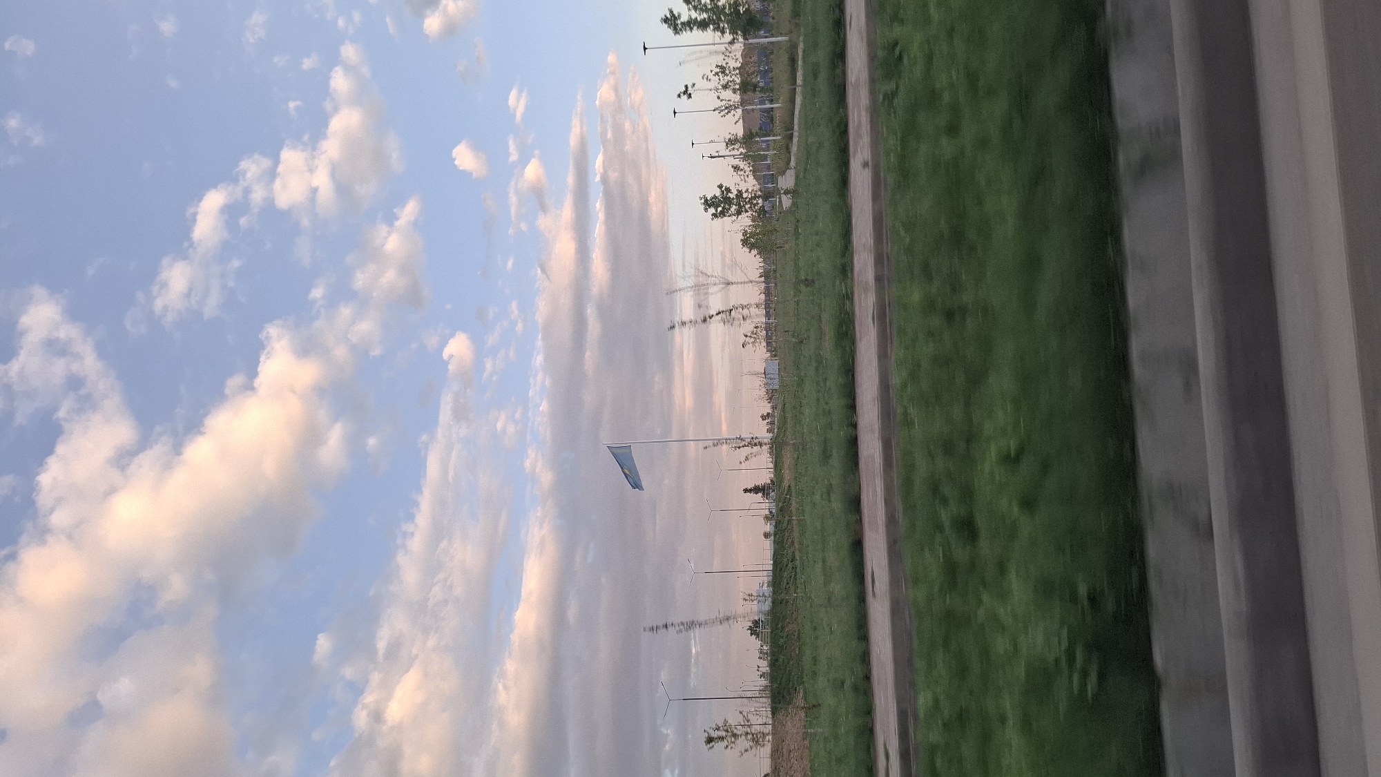 The image depicts a wide, open landscape with a clear sky filled with soft, pink-tinged clouds. A flag, likely the flag of Kazakhstan given the location, is prominently displayed on a tall flagpole in the middle ground. The flag is blue with a golden sun and eagle. Surrounding the flagpole is a grassy area with scattered trees and lampposts, suggesting a park or public space. In the foreground, a blurred out, gray barrier runs horizontally across the bottom of the frame, indicating the photo was taken from a moving vehicle. The overall scene suggests a peaceful, outdoor setting during the late afternoon or early evening, possibly in the city of Shymkent, Kazakhstan, known for its vast open spaces and national symbols.