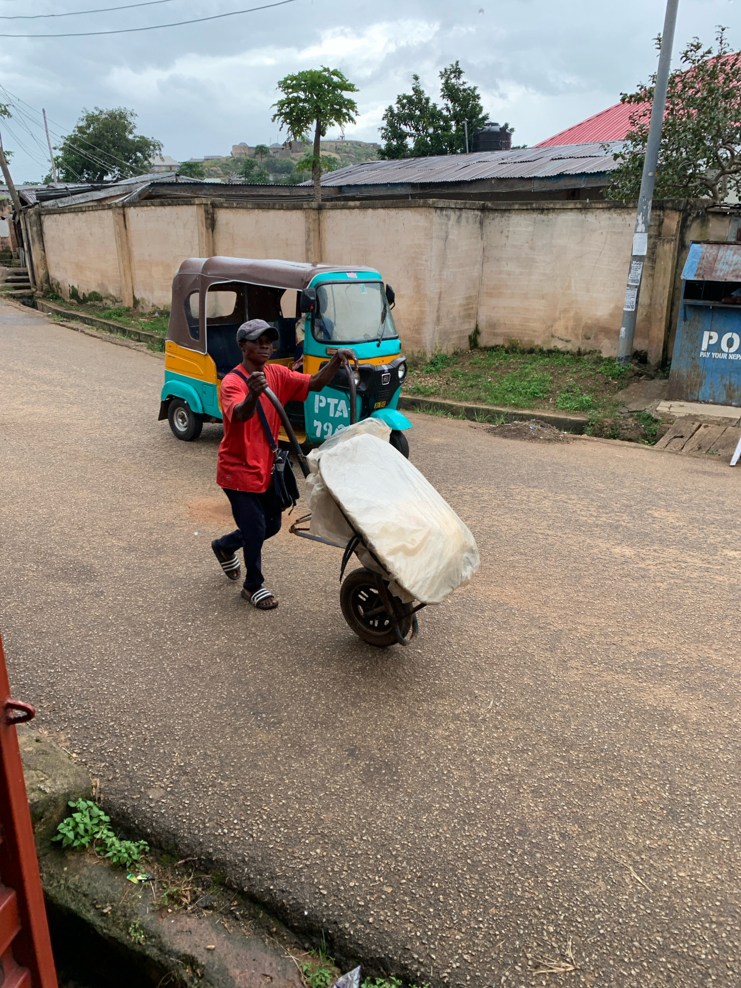 A man, wearing a red t-shirt, dark pants, a baseball cap, and sandals, pushes a wheelbarrow on a paved road. The wheelbarrow is covered with a light-colored tarp. Behind the man is a yellow and teal three-wheeled motorized vehicle, a tuk-tuk, with "PTA 79" visible on its side. The road is bordered by a concrete wall, behind which are buildings with corrugated metal roofs. A utility pole with stickers is also present. The sky is overcast, and vegetation is visible along the side of the road and on a hillside in the distance. The scene appears to be in an urban environment in Jos, Nigeria, likely during a cloudy day.