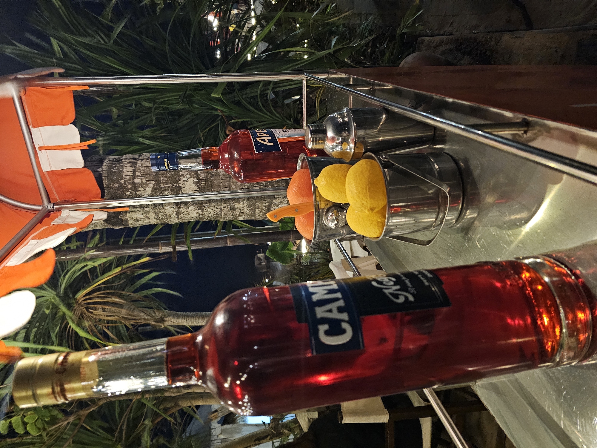 The image depicts a close-up of a bar setup at night, likely in a tropical outdoor setting, given the presence of palm trees and lush foliage in the background. The primary subjects are two bottles of red aperitif and several citrus fruits.
In the foreground, a large, clear glass bottle with a gold cap is tilted to the left. The bottle contains a dark red liquid, and a label with the word "CAMPARI" in blue and white lettering is visible. Below "CAMPARI," the word "Negroni" is partially visible in script.
To the right of the large bottle, a smaller, orange bottle with a blue cap stands upright. Its label also appears to be for an aperitif, with "APEROL" in blue lettering.
Between the two bottles and slightly behind them are two small, silver metal buckets. One bucket contains two whole lemons and the top of an orange with a knife inserted. The other bucket appears to contain additional lemons. A silver cocktail shaker stands to the right of these buckets.
The bar itself is made of a reflective metallic surface, which shows reflections of the bottles and surrounding lights. Above the bar, a canopy with alternating orange and white stripes is visible. The background consists of dark foliage and palm trees, with some out-of-focus lights suggesting a restaurant or event setting.
While the image is presented in the context of Denpasar, Indonesia, there are no explicit visual cues within the image itself to confirm this specific location. The overall scene suggests a beverage service area, potentially for making aperitif-based cocktails. There are no visible people in the immediate frame.