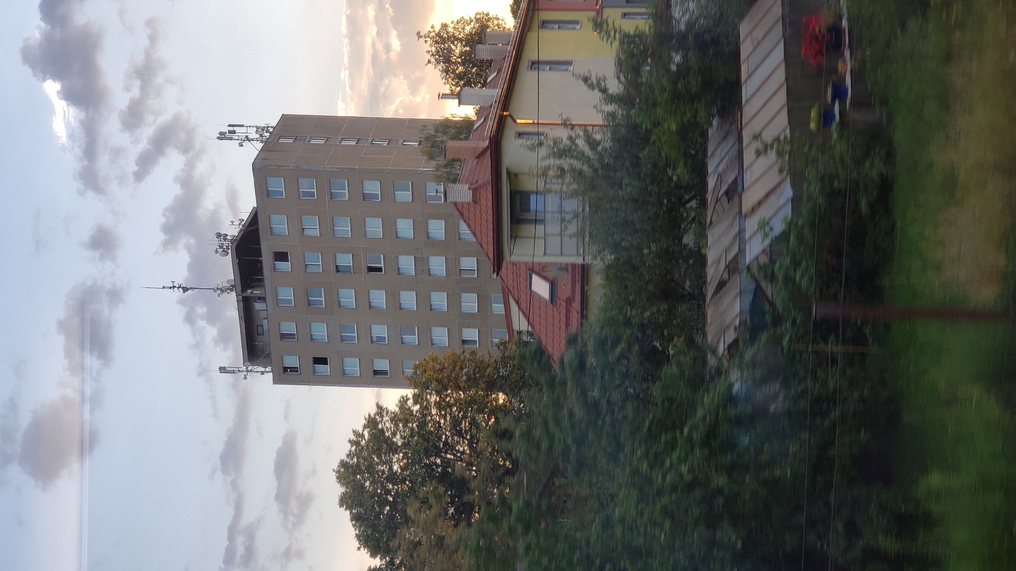 The image shows a tall, concrete apartment building with numerous antennas on its roof, towering over other, smaller buildings and trees. In the foreground, there are green trees and a blurry view of what appears to be greenhouses or industrial sheds. The sky is partly cloudy with a sunset glow. The context suggests the location is the city of Poprad, Slovakia.