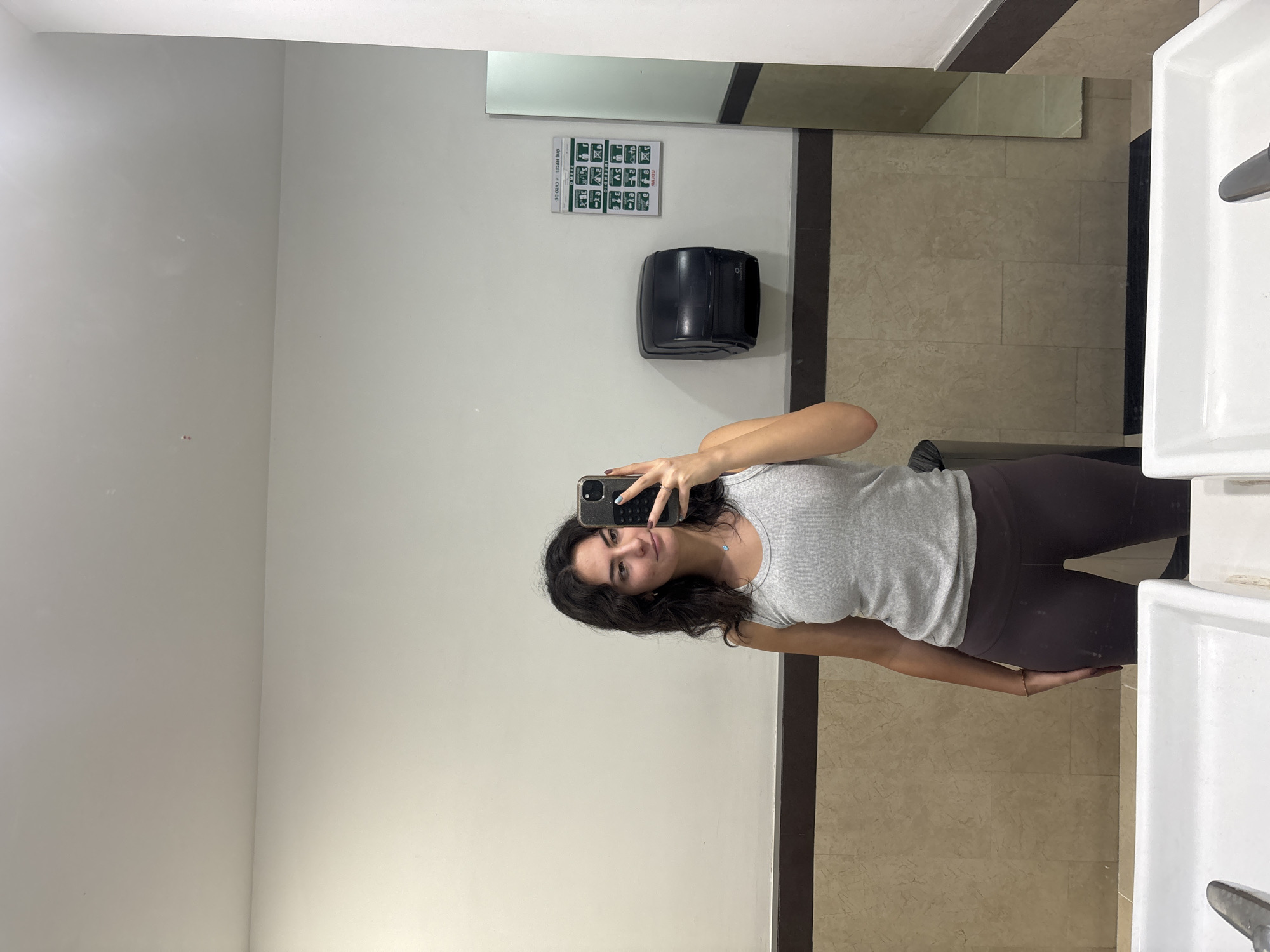 A young woman is captured taking a mirror selfie in what appears to be a public restroom. The image is oriented sideways, showing the woman's reflection along the right side of the frame. She has dark, wavy hair and is wearing a light grey ribbed tank top and dark leggings. She holds a smartphone with a dark case and a small charm, displaying her face with a slight smile. Her fingernails are painted in shades of blue and white.
The background consists of plain white walls and a tiled floor featuring a light brown/beige marble-like pattern. Visible elements in the room's reflection include a black wall-mounted hand dryer and a white rectangular sign with green and black pictorial icons, likely depicting hygiene instructions. Partially legible Spanish text, such as "DUE HACE KYUD DE," is visible on this sign. On the far right, two white ceramic sinks with a chrome faucet are partially captured. The indoor setting, likely in San Andrés Cholula, Mexico, is illuminated by artificial lighting.