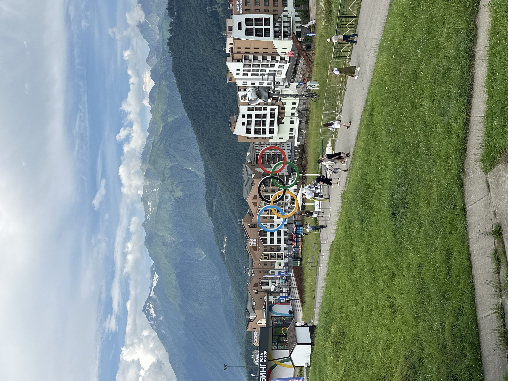 This is a medium shot, eye-level, full outdoor photograph of the Olympic rings monument in Estosadok, Russia, against a backdrop of a village and mountains. The monument, consisting of the five Olympic rings, is prominently featured in the mid-ground. In the foreground, a paved walkway is lined with green grass on the left and a metal fence on the right. People are walking along the paved path. Behind the Olympic rings, a cluster of modern buildings forms a village, with some buildings displaying signage. Beyond the village, lush green mountains rise up, with some of the peaks showing snow or lighter colored rock. The sky is partly cloudy with white clouds scattered across a light blue expanse. The overall scene depicts a scenic mountain resort area on a bright, overcast day.