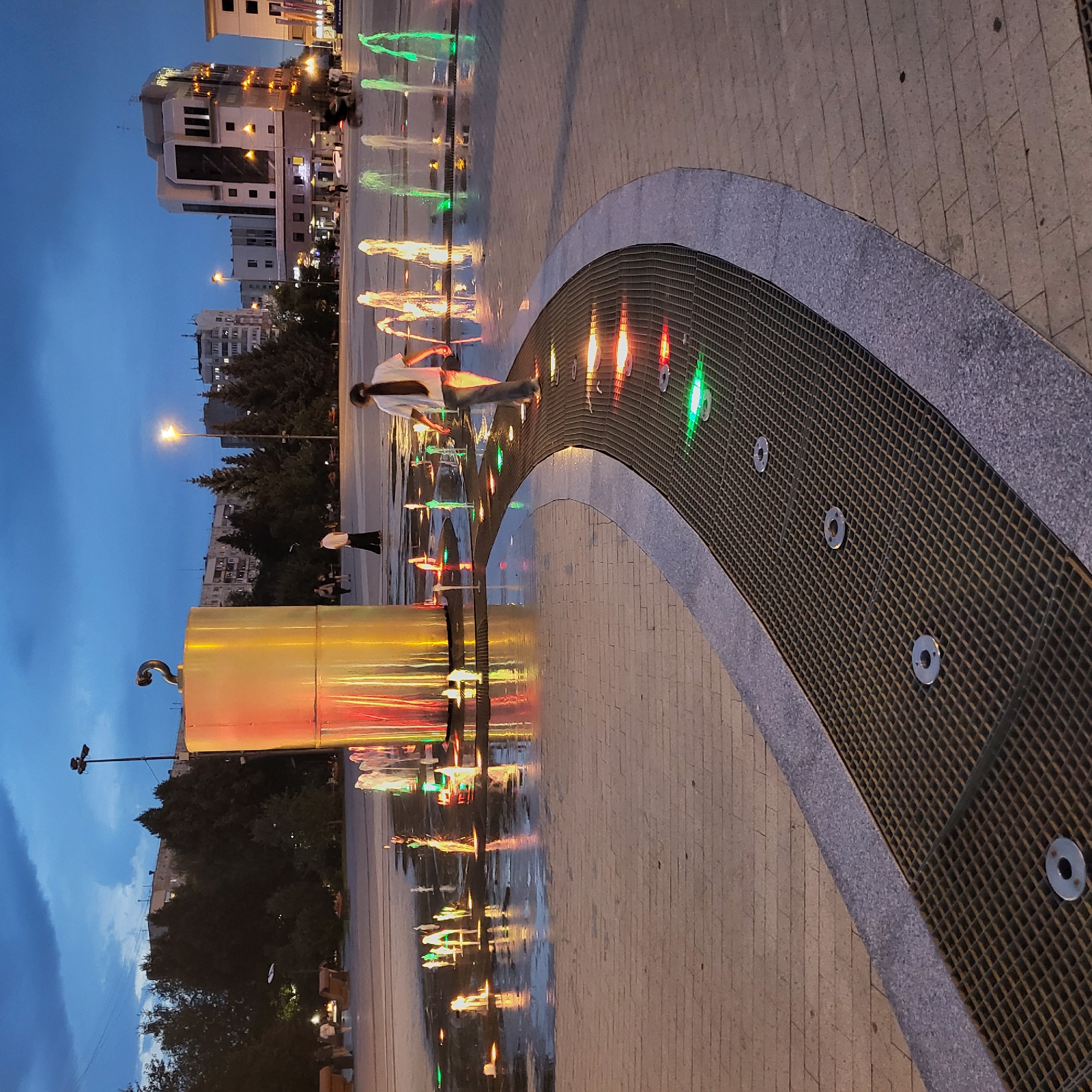 The image depicts an outdoor plaza at dusk in Tyumen, Russia, featuring a modern fountain with illuminated water jets. A large, golden cylindrical structure, possibly a monument or art installation, stands prominently in the background. The plaza is paved with light-colored square tiles, and a curved metal grate with embedded colored lights forms part of the fountain's design. Several people are visible in the scene: a young person with their back to the camera, wearing a white t-shirt and dark pants, is walking along the grate; other individuals are walking in the background, some near benches and trees. The sky is a deep blue, indicating twilight, and buildings of various architectural styles line the perimeter of the plaza. The overall atmosphere is calm and illuminated by the artificial lights of the fountain and surrounding structures.
