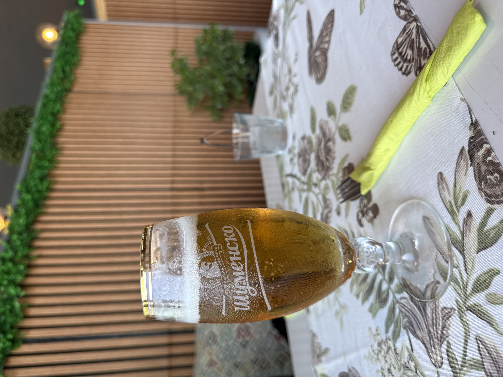 In an outdoor setting, a tall glass of golden beer with a frothy head sits on a white tablecloth adorned with a floral and butterfly pattern. The glass has a gold rim and a logo etched into it that reads "Шуменско" in Cyrillic. To the right of the glass, a fork is wrapped in a bright yellow napkin. In the background, a wall of vertical wooden slats provides a backdrop, with green artificial foliage draped along the top. A small potted plant is visible further in the background, along with a clear glass of water. The overall ambiance suggests a relaxed dining or drinking experience at a restaurant or cafe.
