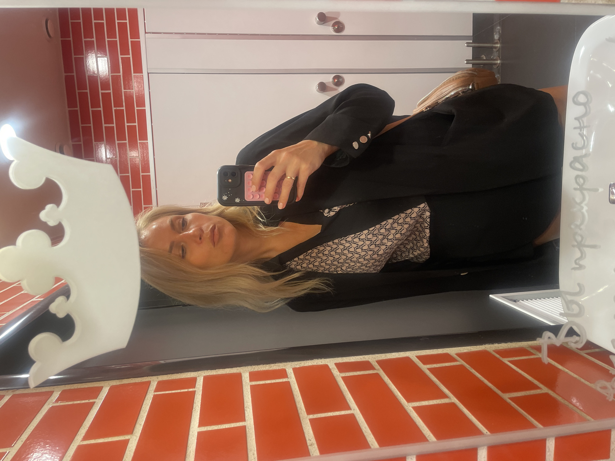 A blonde woman in a blazer is taking a selfie in a bathroom mirror. The bathroom has orange brick tiling and white cabinets with silver knobs. The woman is wearing a patterned top under the blazer. A white crown-shaped object is partially visible in the mirror's reflection, and the word "прекрасно" (beautiful) is written on the mirror's surface. The location is Sochi, Russia.