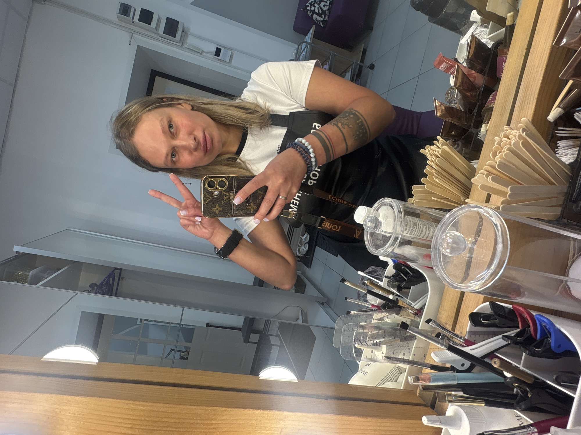 The image is a selfie taken in what appears to be a beauty salon or makeup studio. The location is likely Perm, Russia, as indicated by the "TORRAS" branding visible on the apron worn by the person in the image, which is a well-known cosmetic brand in Russia.
The subject of the photo is a fair-skinned woman with blonde hair, wearing a white t-shirt and a black apron. She has tattoos on her left forearm and is wearing bracelets and a watch on her left wrist. She is holding a smartphone with a Louis Vuitton patterned case, taking the selfie in a mirror. She is making a peace sign with her fingers.
In the background, there is a table with various makeup tools and supplies. These include makeup brushes, containers of makeup applicators, and packaging of cosmetic products. There is also a shelf visible in the background, and possibly a purple chair. The lighting in the salon is bright, likely due to the illuminated mirror.