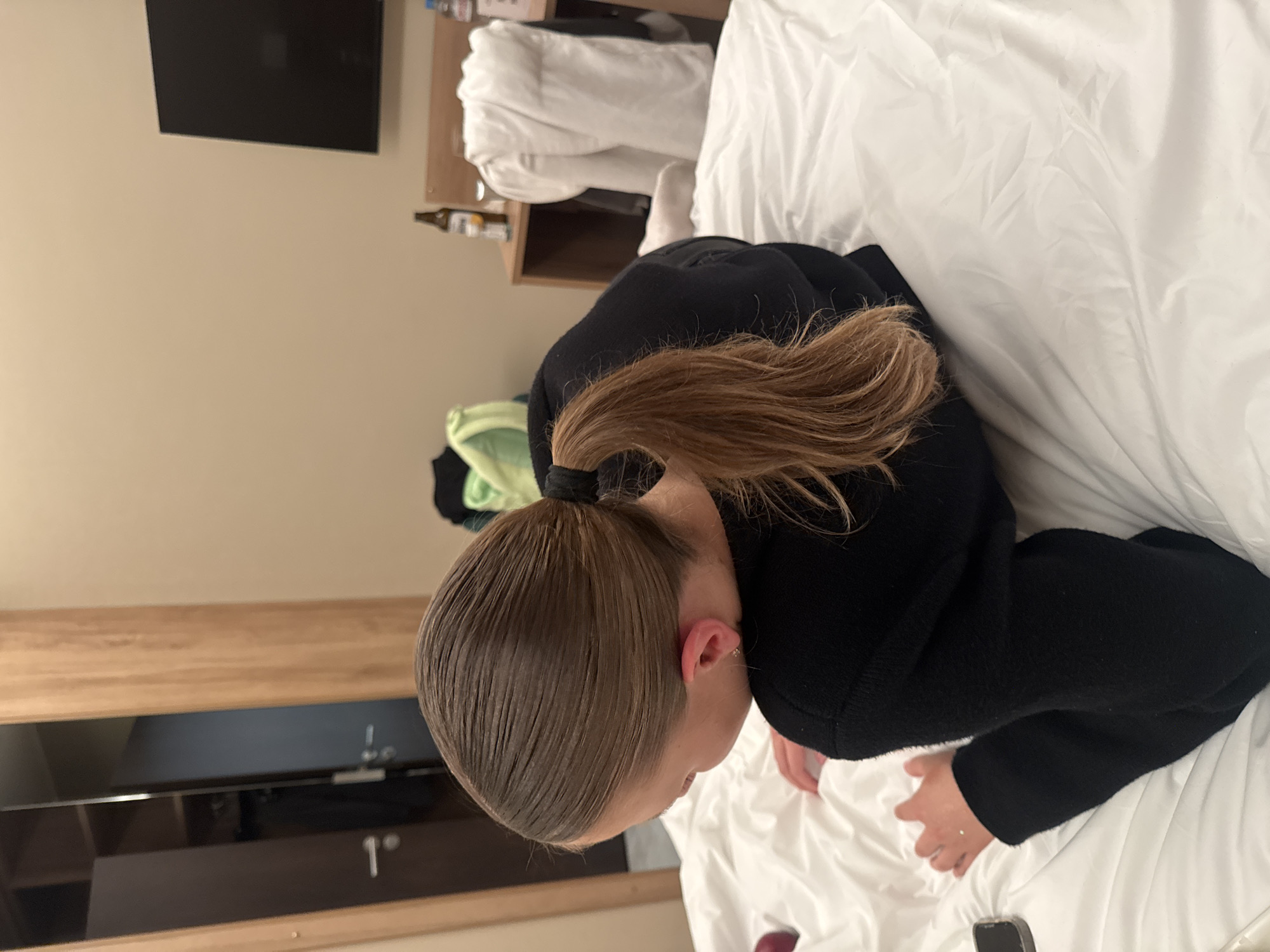 A young person with blonde hair pulled into a ponytail is lying on a bed covered with white sheets. The person is wearing a black top. The room appears to be a hotel room with a closet visible in the background to the left, and a bedside table with a television and some bottles to the right. The lighting is dim, suggesting it might be evening or nighttime.