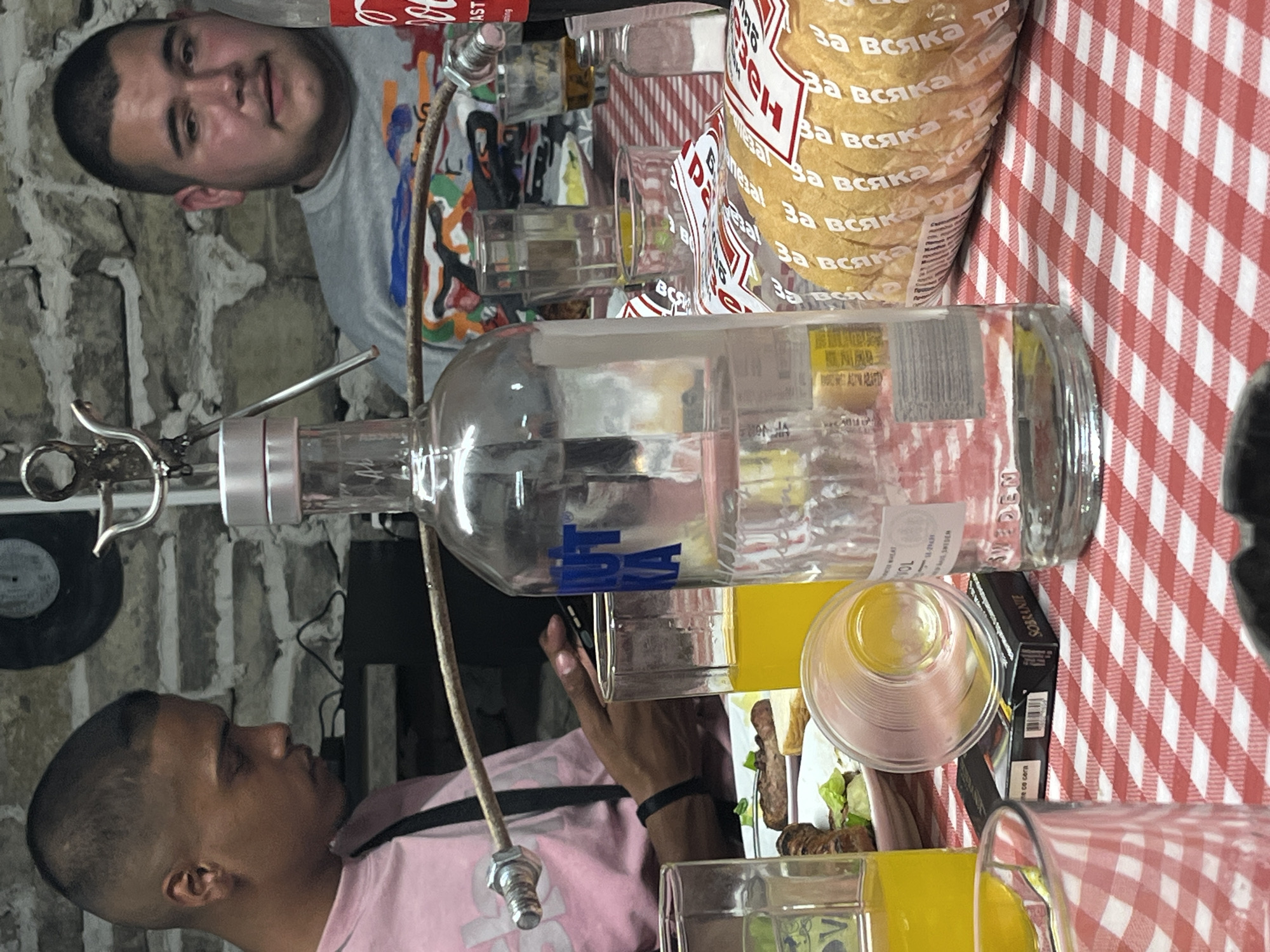 The image shows two men at a table covered with a red and white checkered tablecloth. A bottle of Absolut vodka is prominently displayed. A package of "За всяка ТР" (Za vsyaka TR) snack food is also on the table. There are drinks in glasses, and a pack of Sobranie cigarettes near a plate with what appears to be a salad or side dish. The men seem to be seated in an indoor environment with stone walls. The location appears to be Lozarevo, Bulgaria, based on the Cyrillic script on the package.