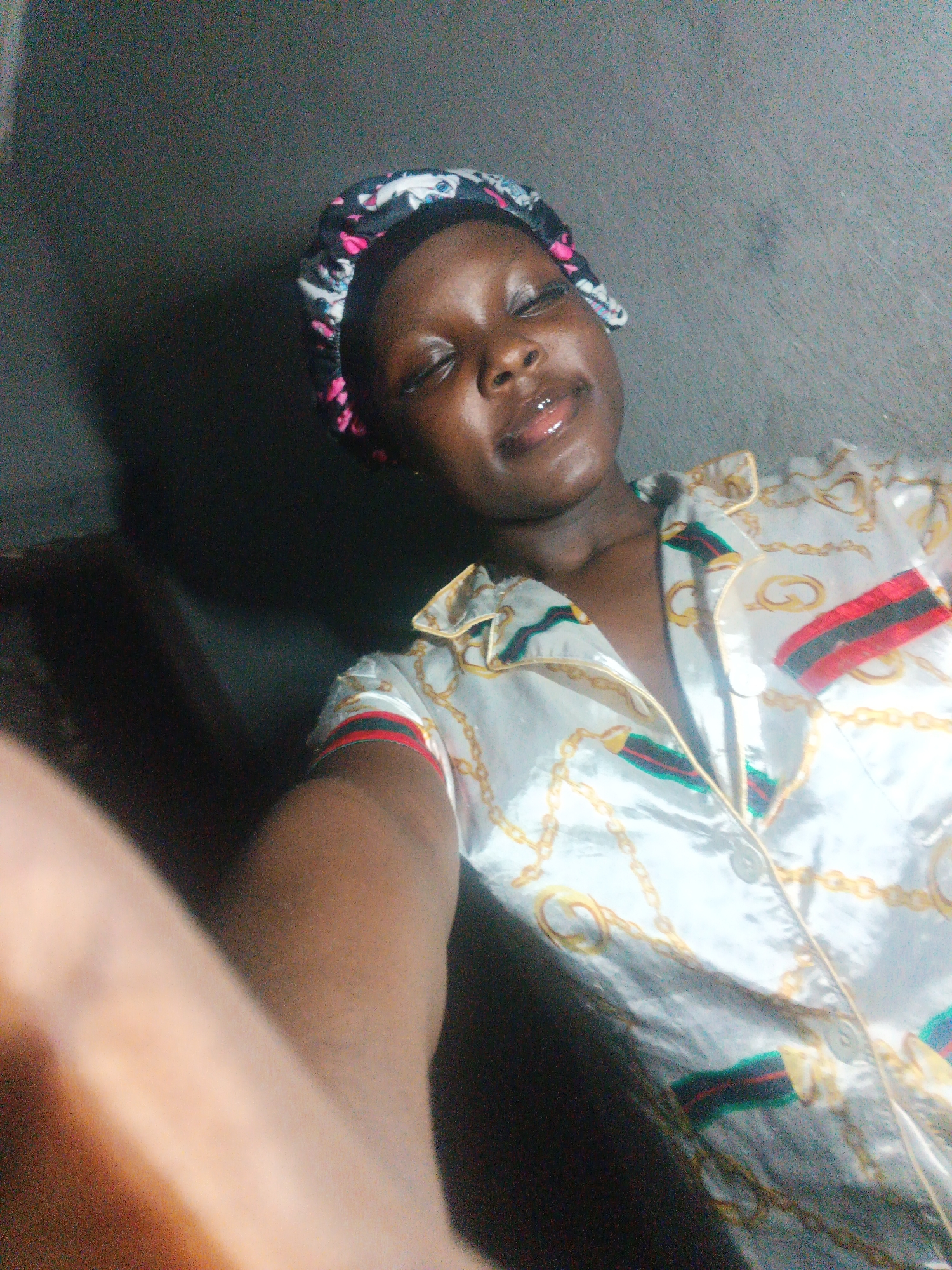 This selfie captures a young Black person indoors, likely in Onitsha, Nigeria. The individual, appearing female, is seen from the chest up, with their head tilted slightly. Their eyes are closed or squinting, and they have a gentle smile, suggesting a relaxed or happy mood. Their lips appear glossy.
They are wearing a multi-colored sleep bonnet or head covering, featuring black, pink, white, and blue patterns. Over their body, they wear a shiny, light-colored pajama top, possibly satin, adorned with intricate gold chain designs and distinctive red, green, and black striped accents on the collar and sleeves.
A forearm and hand are visible in the lower-left foreground, indicating the self-portrait nature of the image. The background is a dark, plain, textured wall, possibly concrete. The lighting is somewhat dim but illuminates the subject's face. There is no visible text in the image.