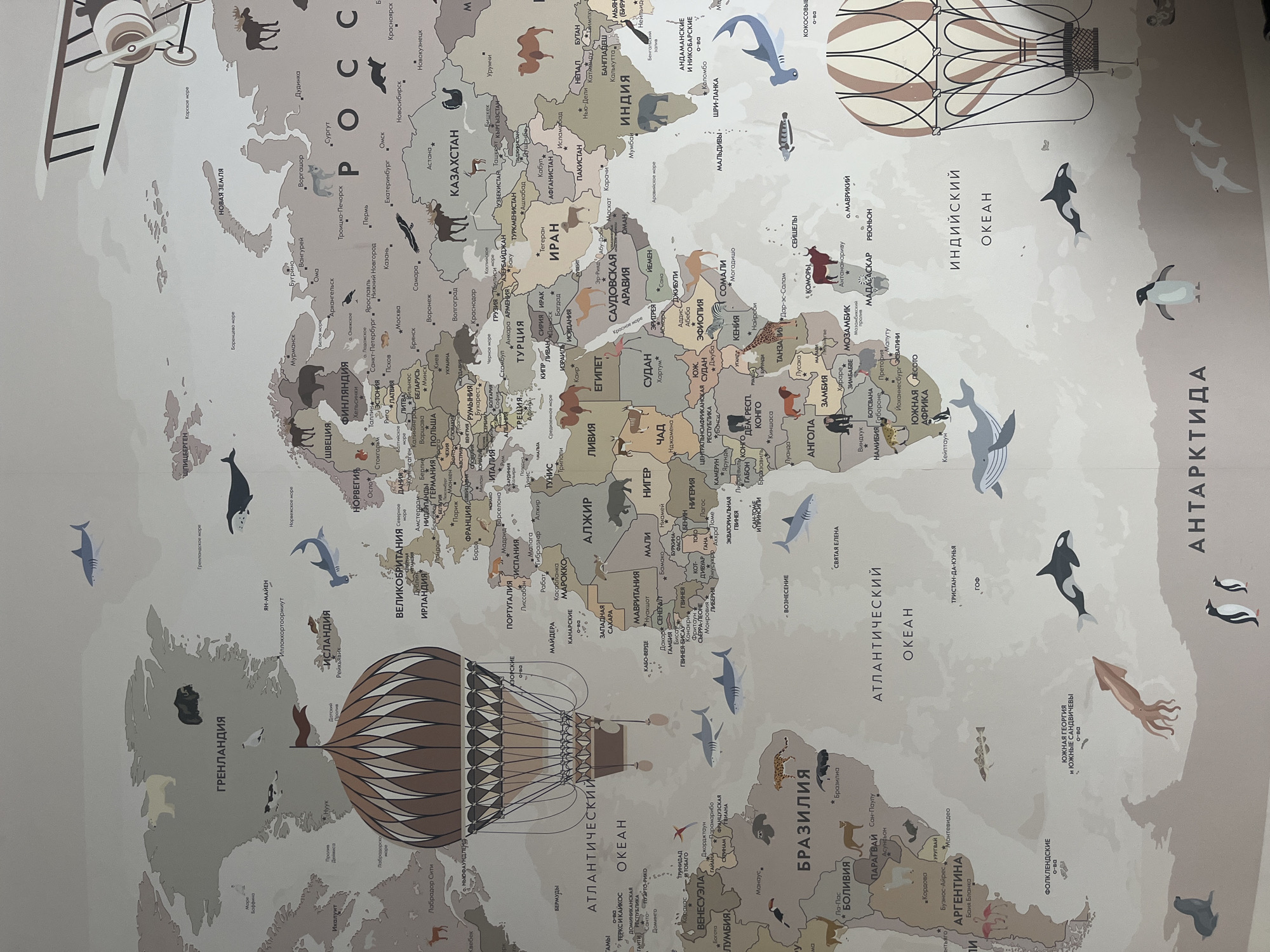 The image shows a detailed map of the world, illustrated with animals and geographical features. It includes countries like Russia, Kazakhstan, India, Brazil, and Argentina. Different oceans, such as the Atlantic, Indian, and Arctic, are labeled, along with locations like Greenland and Antarctica. The map is a light beige color with varied shades of brown and grey used to define continents and countries. There are illustrations of hot air balloons. The city of Kaspiysk, Russia is not visible.
