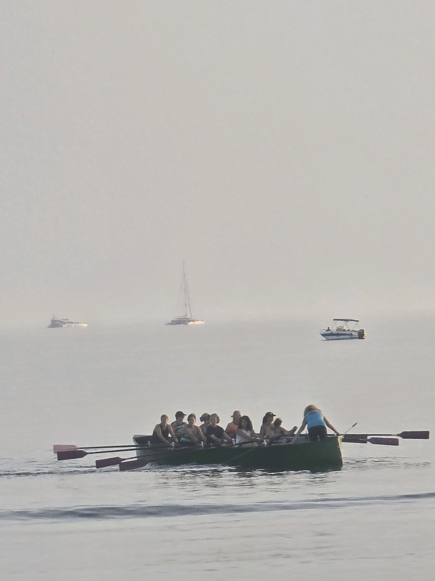 In the image, a group of eight people are rowing a long, dark green boat on calm, misty water. Seven individuals are seated inside the boat, actively rowing with oars. One person, a woman wearing a blue top and dark pants, is standing at the stern of the boat, facing away from the camera and holding an oar. The boat is moving from left to right, creating a visible wake. In the background, there are at least three other vessels visible through the fog: a catamaran sailboat to the left, a larger boat or ship in the middle distance, and a small motorboat with a canopy to the right. The environment appears to be an open body of water, possibly the sea or a large lake, with a very hazy or foggy atmosphere that obscures the horizon. The overall lighting suggests it is either early morning or late evening. Without additional information, the specific location in Málaga, Spain, cannot be determined from the visual data alone.