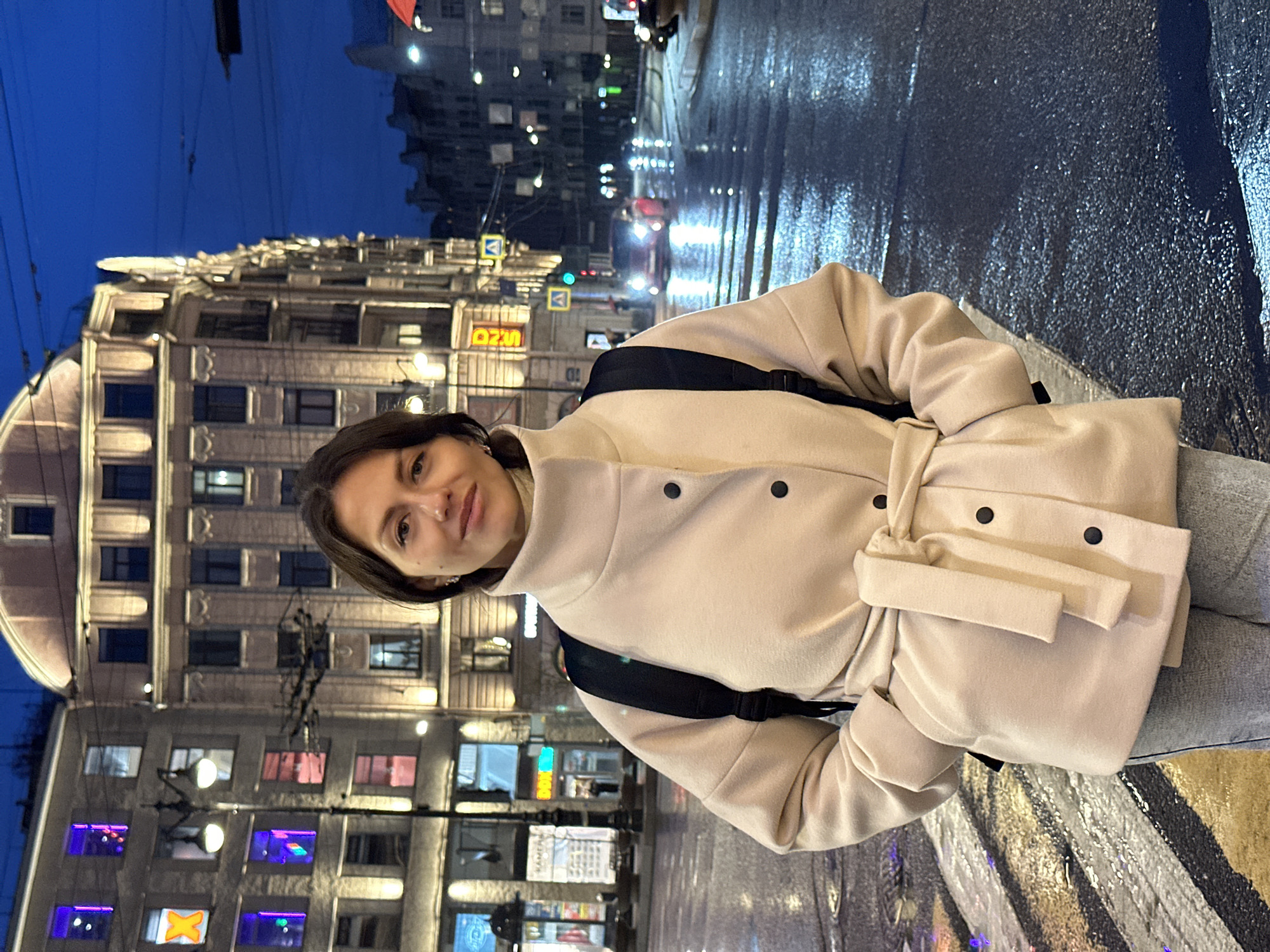 A medium-shot, rotated, ground-level photograph taken at night depicts a woman, likely Caucasian, standing on a wet street in Saint Petersburg, Russia. The woman is wearing a light beige, belted coat and a black backpack. The buildings in the background are illuminated with streetlights and neon signs. The street is wet, reflecting the lights of the city. The style of the buildings suggests the location could be in the city center, known for its historical architecture. The sky is a dark blue hue. The image appears to be taken with a smartphone, with the lens close to the subject and focusing on the foreground.