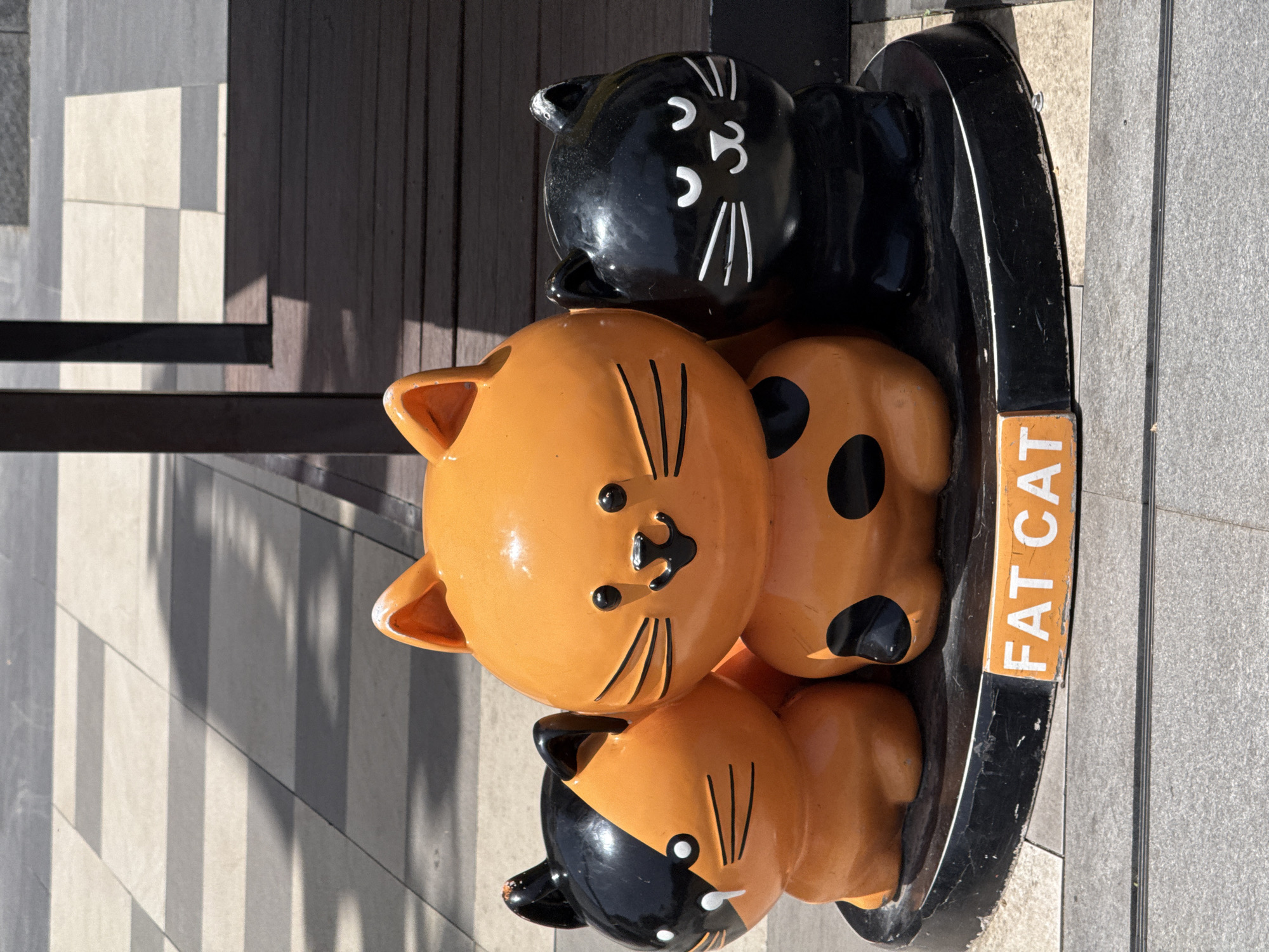 The image shows a sculpture of three stacked ceramic cats in Yanta District, China. The bottom cat is orange and black, the middle cat is orange with black spots, and the top cat is black with white markings. The sculpture is situated on a black base labeled "FAT CAT" in orange letters. The background features a boardwalk and concrete paving.