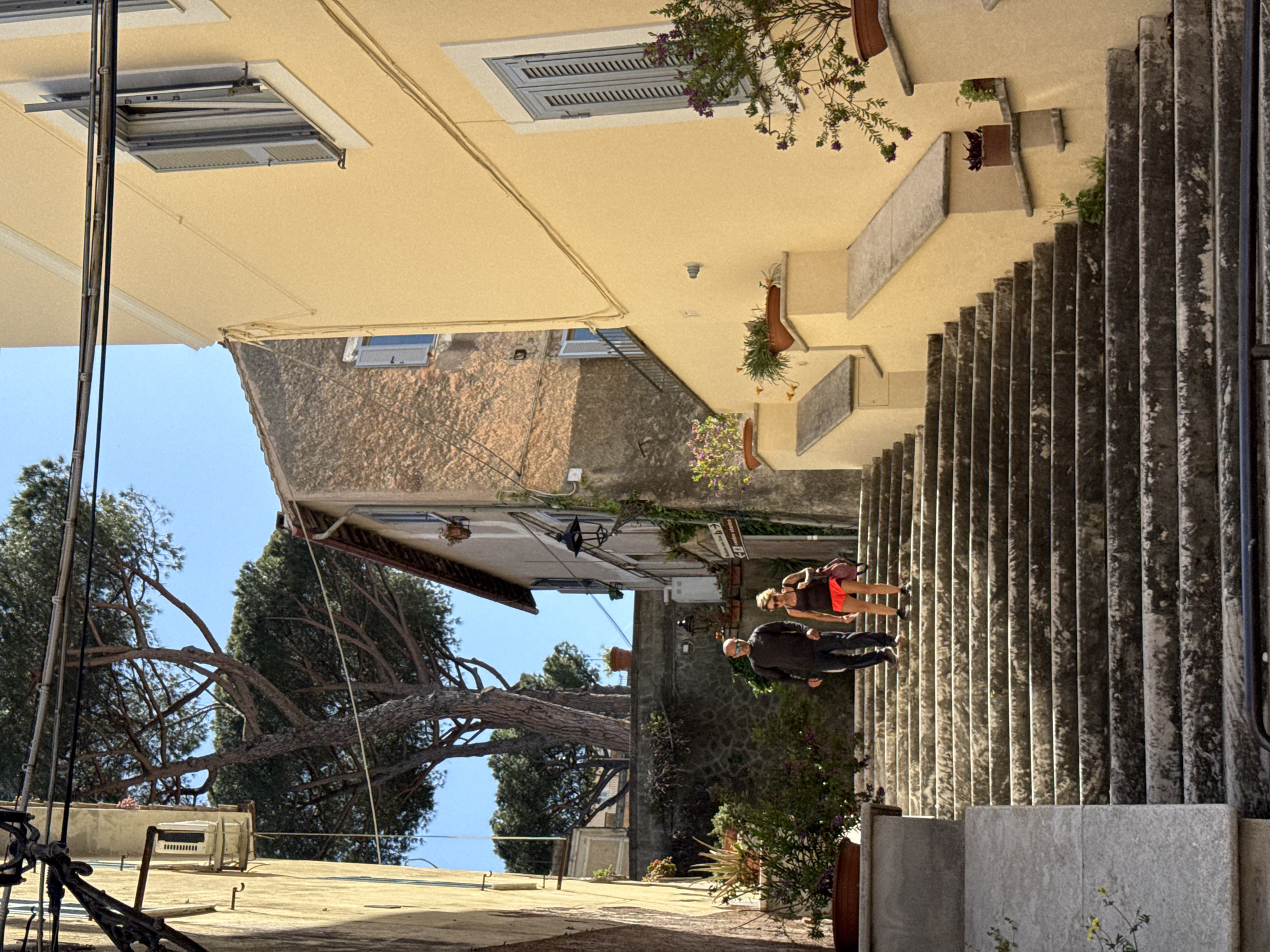 The image captures a sunlit, stepped street or pathway in Castel Gandolfo, Italy, presenting a picturesque scene of traditional architecture and natural elements. On the right, a wide stone staircase descends, bordered by light yellow-painted buildings. These buildings feature windows with closed light grey shutters and are decorated with numerous potted plants, some with purple flowers, on ledges and along the steps.
Two individuals are visible on the upper part of the stone steps, appearing to walk downwards. The person in front is a man, wearing a dark jacket and trousers, with a light-colored face mask visible. Behind him is a woman, dressed in a dark top and reddish-orange skirt or shorts, carrying a backpack.
To the left of the pathway, a large, gnarled pine tree with expansive branches frames the view against a clear blue sky. Further down this side, another building with a textured, rustic facade and shuttered windows can be seen.
The bright sunlight casts distinct shadows, indicating a clear, sunny day, likely mid-morning or afternoon. Visible details include an air conditioning unit on a distant rooftop and a black lamppost. A white street sign mounted on a stone wall clearly displays the text "L.go L.go Umberto I". The scene conveys a sense of peaceful ambiance within a historic Italian town.