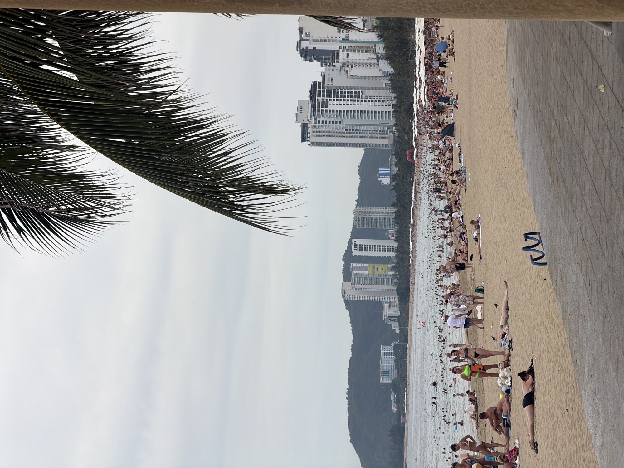 The image captures a bustling beach scene in Nha Trang, Vietnam. In the foreground, a concrete walkway is visible, with a rough, textured surface. To the right, a section of a cylindrical, light-brown pillar partially obstructs the view. The sand beach is populated with numerous individuals sunbathing, strolling, and interacting. Some are lying on towels, while others are standing or walking along the shore. A group of people are clustered closer to the camera, with some wearing swimwear. Further back, the ocean is filled with swimmers and various flotation devices. Along the shoreline, a long stretch of umbrellas and beach chairs are set up, indicating a popular tourist area. In the background, a densely populated coastline is dotted with palm trees and a significant number of high-rise buildings, characteristic of a city. The buildings vary in height and architectural style, with some appearing modern and white. Beyond the city, a large, green, mountainous landscape forms the backdrop under an overcast sky. The overall impression is one of a vibrant, active beach resort on a cloudy day.