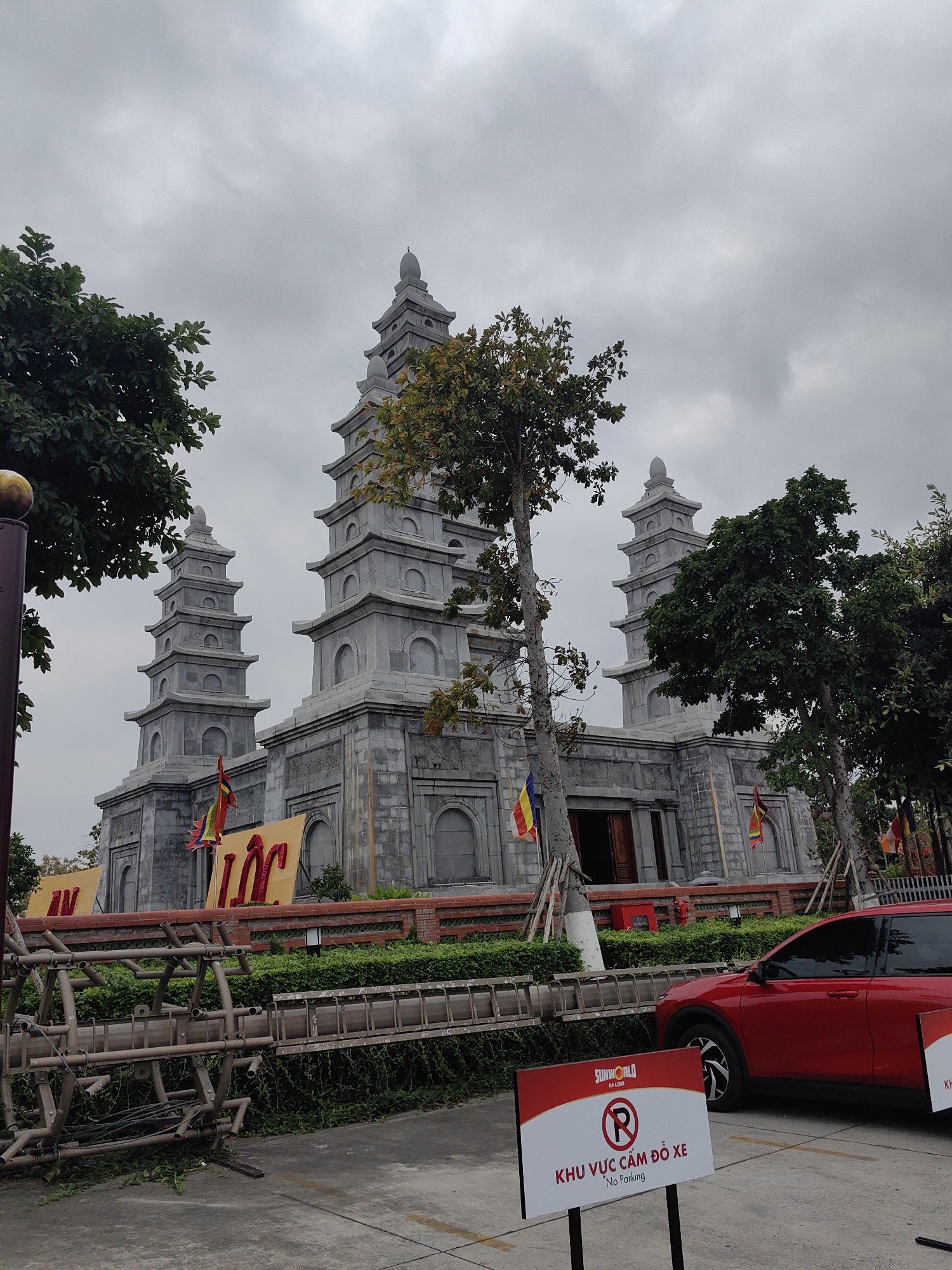 The image presents a large, elaborate stone building featuring multi-tiered pagoda-style architecture under an overcast sky. The structure is built from grey stone, with several towers of varying heights, each characterized by multiple stacked levels and arched recesses. Green trees and dense foliage surround the building, partially framing it.
In the midground, a low red brick wall runs along the base of the building, behind which large yellow banners with prominent red Vietnamese characters, including "LỘC," are displayed. Colorful flags, resembling Buddhist prayer flags, are visible near the building's entrances.
The foreground includes a grassy area with additional trees and a significant amount of metal scaffolding or framing on the ground to the left. To the right, the front portion of a red SUV is visible. A red and white sign stands in the lower right corner, bearing the text "SUNWORLD HA LONG" at the top, a no-parking symbol, and the phrases "KHU VỰC CẤM ĐỖ XE" and "No Parking" in both Vietnamese and English.
The weather is cloudy and overcast, suggesting a daytime scene without direct sunlight. No people are visible in the photograph, contributing to a calm and static atmosphere. The visible text "Sun World Ha Long" provides a clear location cue, placing the scene in a tourist complex in Hong Gai Ward, Vietnam.