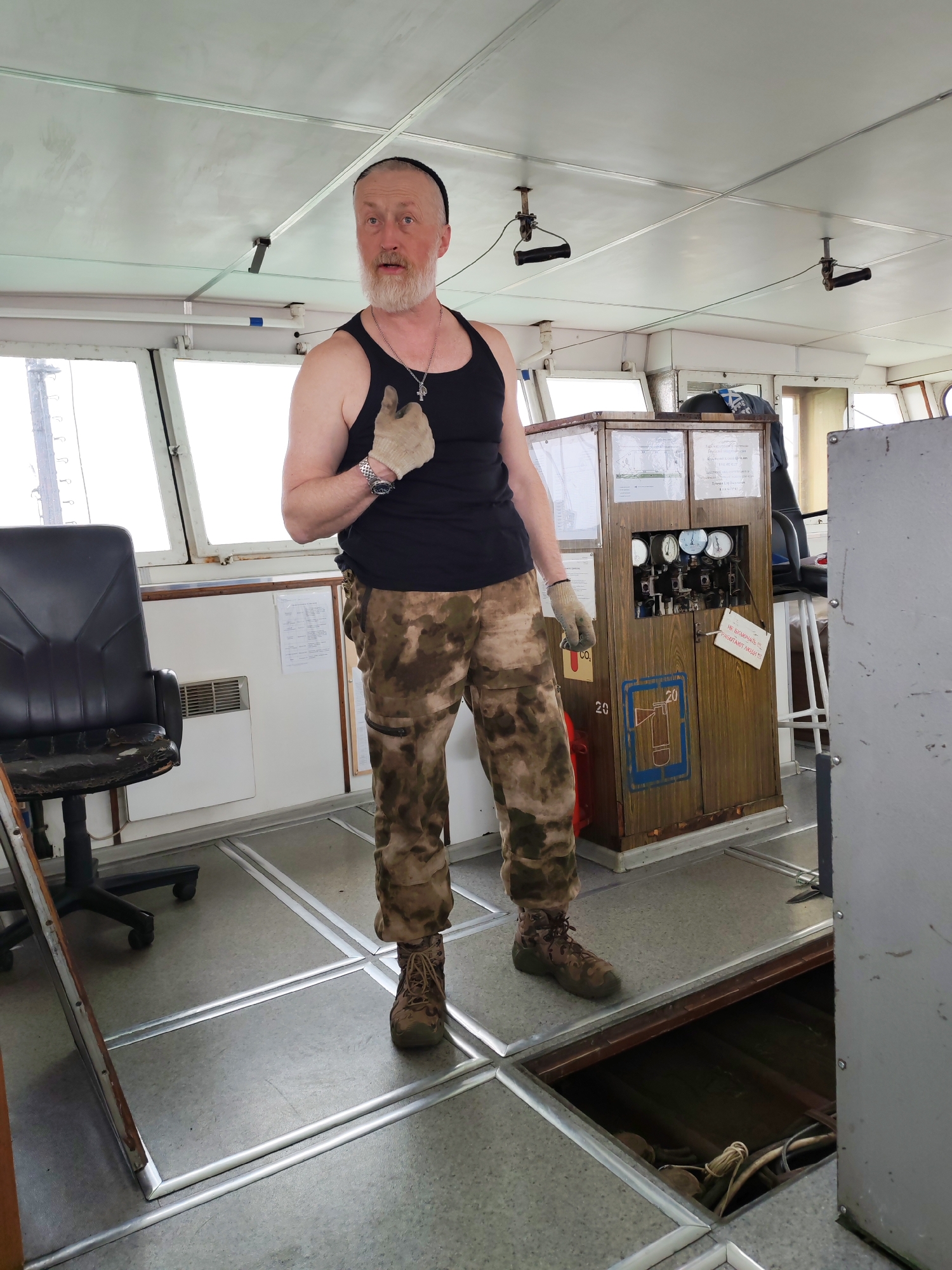 A man with a white beard, wearing a black tank top, camouflage trousers, and work gloves, stands on what appears to be the bridge or control room of a vessel, likely in Shcheleiki, Russia. He has a black cap on his head, a silver chain with a pendant around his neck, and a watch on his left wrist. His mouth is slightly agape, and he gives a thumbs-up gesture with his right hand, looking directly at the camera.
The interior features large windows offering a bright, possibly overcast, view outside. The floor is covered with gray material divided by silver metal strips. To the man's left is a worn black office chair. To his right, a tall, wooden cabinet serves as a control panel, fitted with several gauges and switches. A red "CO2" label and a blue stencil of a fire extinguisher with the number "20" are visible on its lower half. A white sign hanging from the cabinet reads, "НЕ ВКЛЮЧАТЬ! НА РАБОТАЮТ ЛЮДИ!" (DO NOT SWITCH ON! PEOPLE ARE WORKING!). An open hatch in the floor to the right reveals ropes and cables below deck. The ceiling is paneled and has handles hanging down, typical of a marine environment. The overall impression is an operational but somewhat aged maritime workspace.