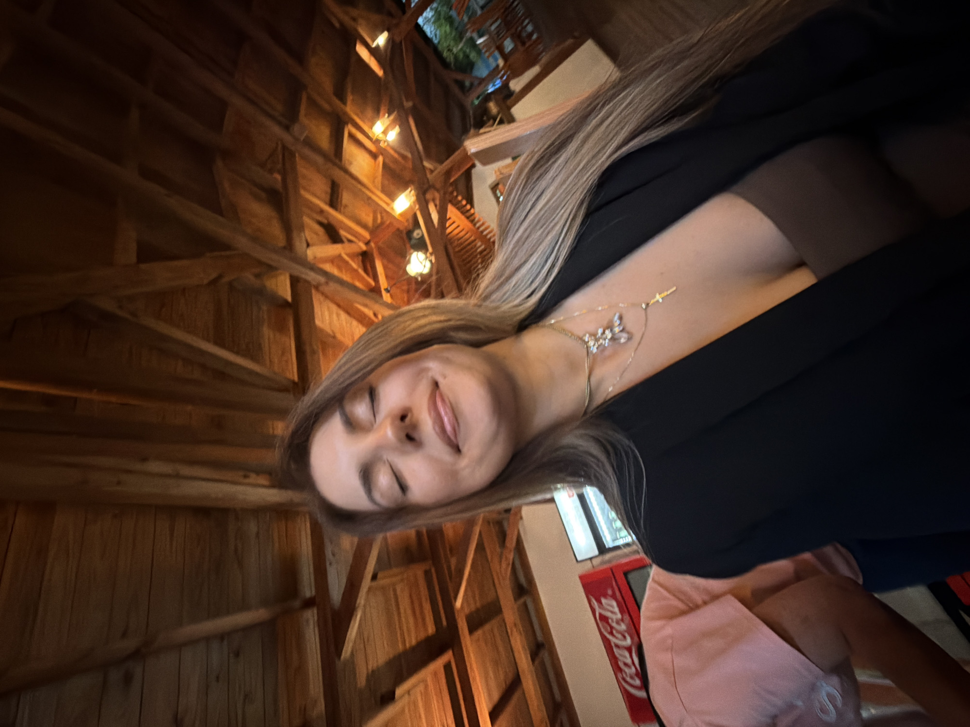This media file shows a woman in the foreground, with her head tilted to the side and eyes closed, appearing to be smiling. She has long blonde hair and is wearing a black jacket over a pink shirt. A delicate silver necklace with a butterfly pendant is visible around her neck.
The background features a rustic interior with exposed wooden beams and a high ceiling, suggesting a traditional or lodge-like setting. Several light fixtures are visible along the beams, emitting a warm glow. In the distance, there's a glimpse of what appears to be a Coca-Cola branded vending machine or sign, indicating a commercial space. The lighting suggests it is either evening or an indoor setting with artificial light. The overall atmosphere seems relaxed and casual. The scene is likely indoors, possibly a restaurant or a similar public venue, given the presence of the Coca-Cola branding. The woman's closed eyes and smile convey a sense of contentment or enjoyment.
