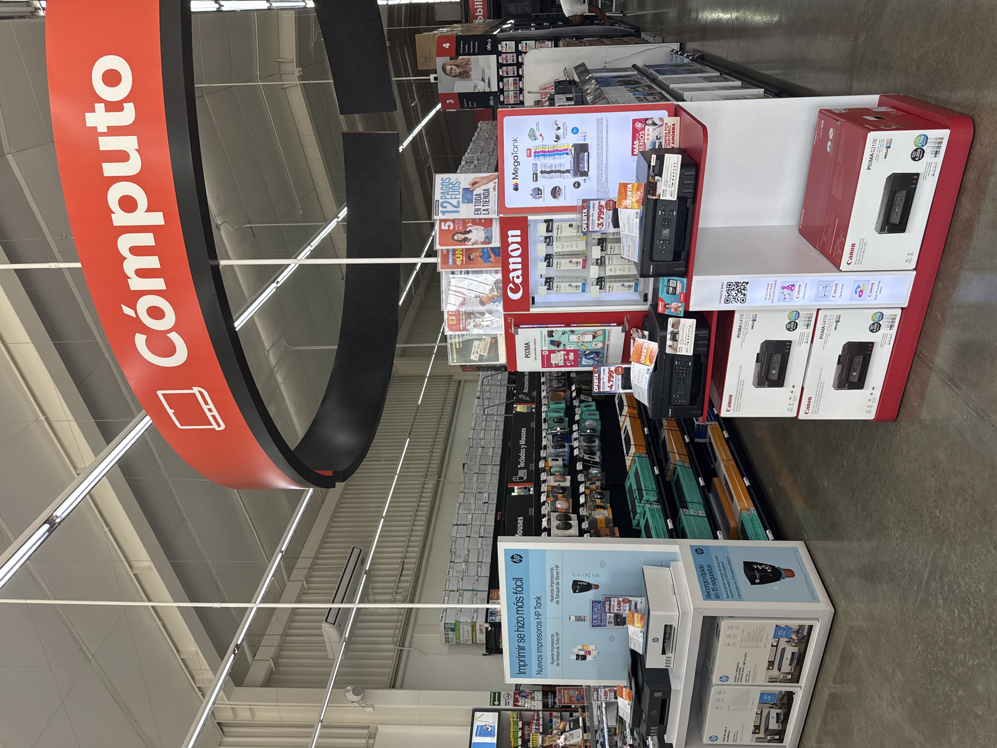 The image captures an interior view of an electronics or office supply store aisle, likely in San Bernardino Tlaxcalancingo, Mexico, given the provided context and Spanish signage. The scene is brightly lit by fluorescent lights and appears well-organized.
The main subjects are various electronic products and display stands. Prominently featured are printers from brands like Canon (MegaTank, PIXMA G4170, PIXMA G2170) and HP (HP Tank series), displayed on multi-tiered stands with accompanying advertisements and product boxes. Boxes for Canon printers are stacked on shelves. Further down the aisle, shelves are stocked with computer accessories, labeled "Teclados y Mouses" (Keyboards and Mouses). A partial view of a person, possibly a customer or employee, is visible in the upper right background.
The activity taking place is typical retail display and shopping. The store is set up to showcase products and inform customers about offers.
Notable details include a large, red, and black circular overhead sign with the word "Cómputo" (Computers) and a laptop/tablet icon, indicating the section of the store. Numerous advertisements are visible, promoting payment plans such as "12 PAGOS FIJOS" (12 Fixed Payments) across "TODA LA TIENDA" (the entire store) and "OFERTA" (Offer) banners with prices like "$3,799" and "$4,799". Other signs highlight features like "Recarga rápida en 15 segundos" (Fast refill in 15 seconds) for HP printers and "COMPRA POR MAYOREO Y AHORRA" (Buy Wholesale and Save) for Canon products. The "Ofix" brand is also visible on several elements. The store has a modern, industrial feel with an exposed ceiling structure.