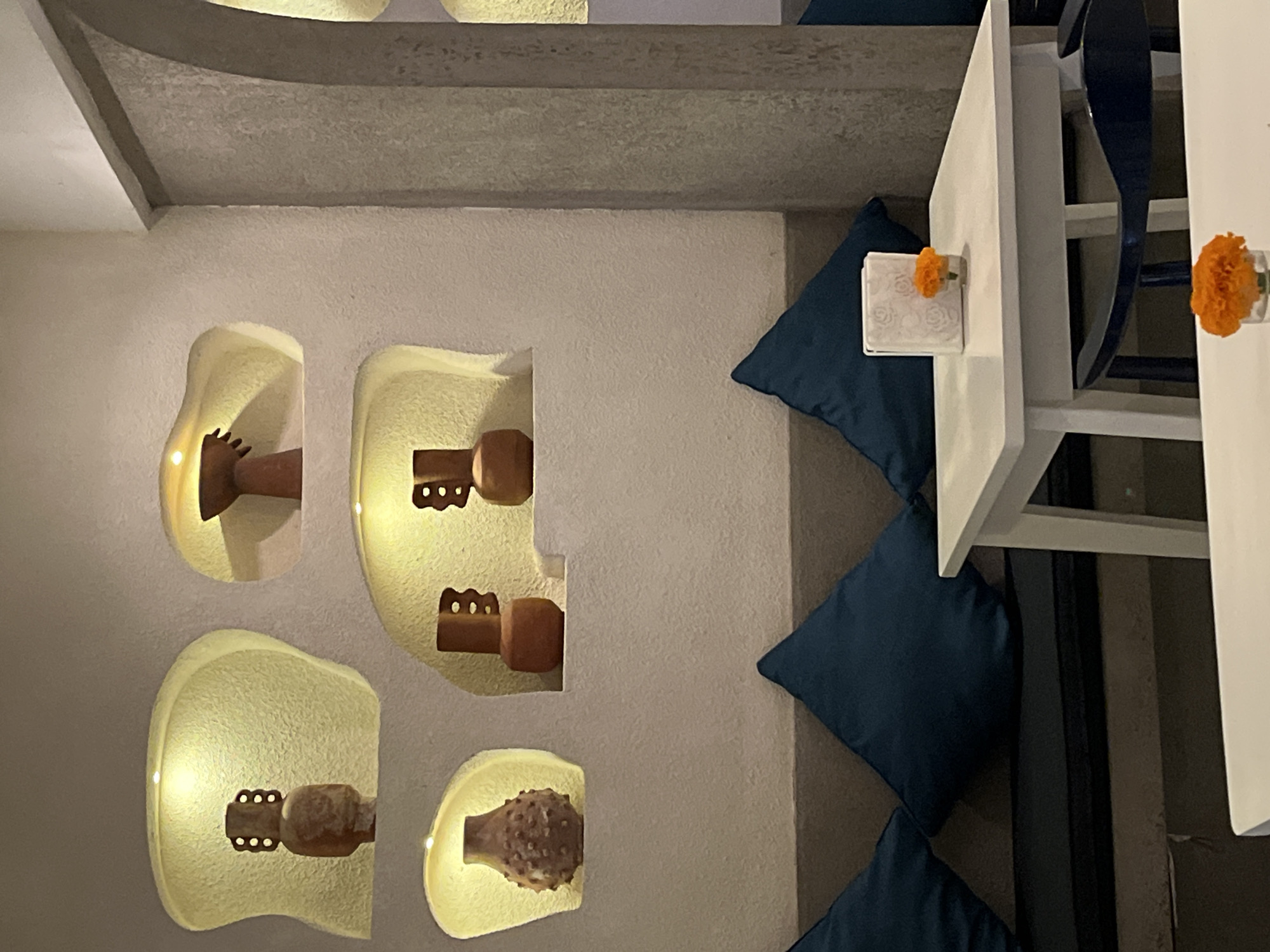 The image depicts the interior of a restaurant, presumably in Unknown, Indonesia, showcasing a unique seating arrangement with blue cushions stacked as a base for patrons. The seating area is adjacent to a wall adorned with recessed niches containing decorative wooden artifacts. A low white table with a small flower placed on top suggests a relaxed and inviting ambiance. The image, captured in an indoor setting, focuses on the aesthetic and functional aspects of the restaurant's design, highlighting the cultural and artistic elements of the decor.
