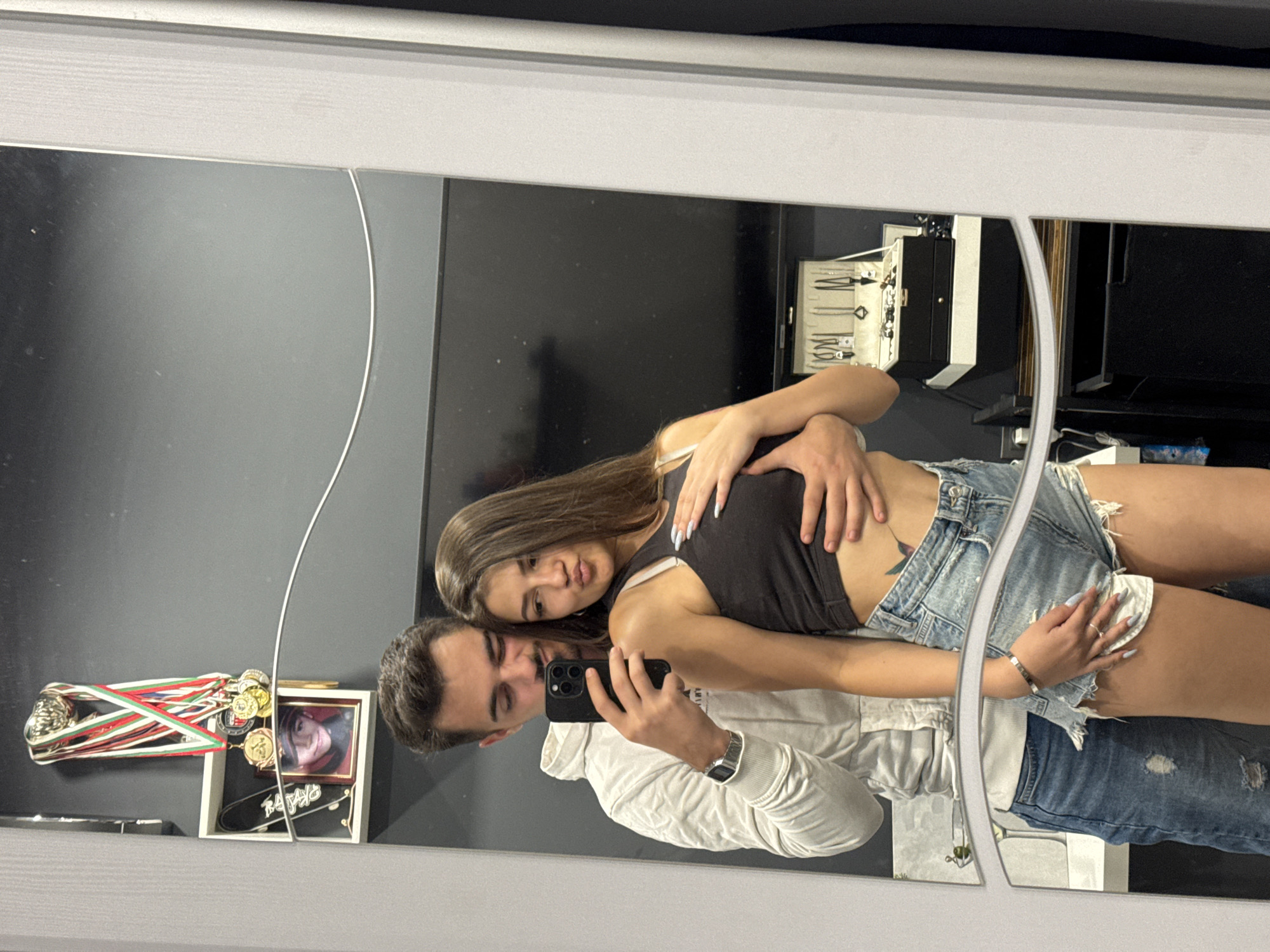 A young couple, a man and a woman, pose for a mirror selfie in what appears to be a bedroom or dressing area, likely in Sofia, Bulgaria. The man, wearing a white hoodie and blue jeans, holds a smartphone to take the picture, with his arm around the woman. The woman, in a black crop top and denim shorts, leans into him. In the background, a shelf displays trophies and medals, a framed picture of a young boy, and a skateboard. A television is mounted on the wall, and a jewelry box sits on a shelf below. The room is dimly lit, with a dark gray wall.