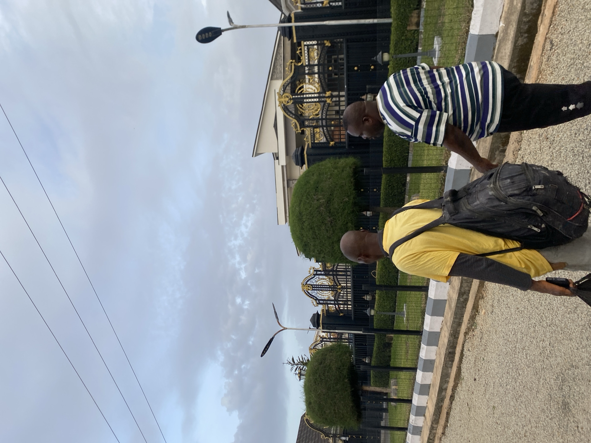 Two men are walking on a gravel road in front of a large, ornate black and gold gate that leads to a modern house. The man on the left wears a yellow t-shirt and a black backpack, while the man on the right wears a striped t-shirt and black pants. The sky is overcast with some visible clouds. The scene appears to be in Barikin Jirga Summa, Nigeria.