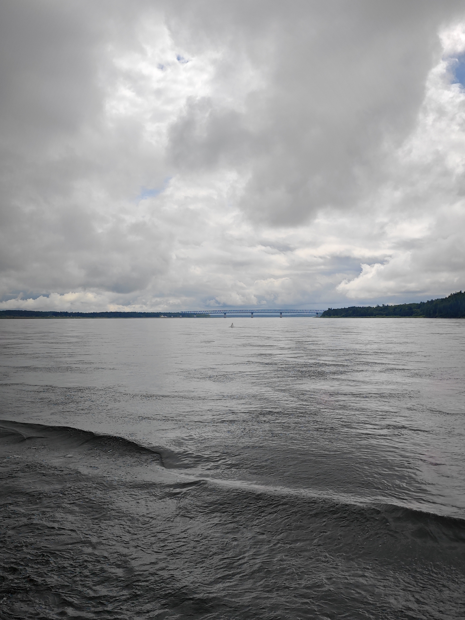 The image depicts a wide river under a cloudy sky. In the distance, a long bridge spans the water, connecting the two shores. The riverbanks are lined with dense green trees. The water in the foreground is dark and choppy, with visible ripples and waves. The sky is filled with heavy, grey clouds, with only small patches of blue sky visible. The scene appears to be a natural landscape with man-made structures. The location is specified as Vysokogorsky District, Russia.