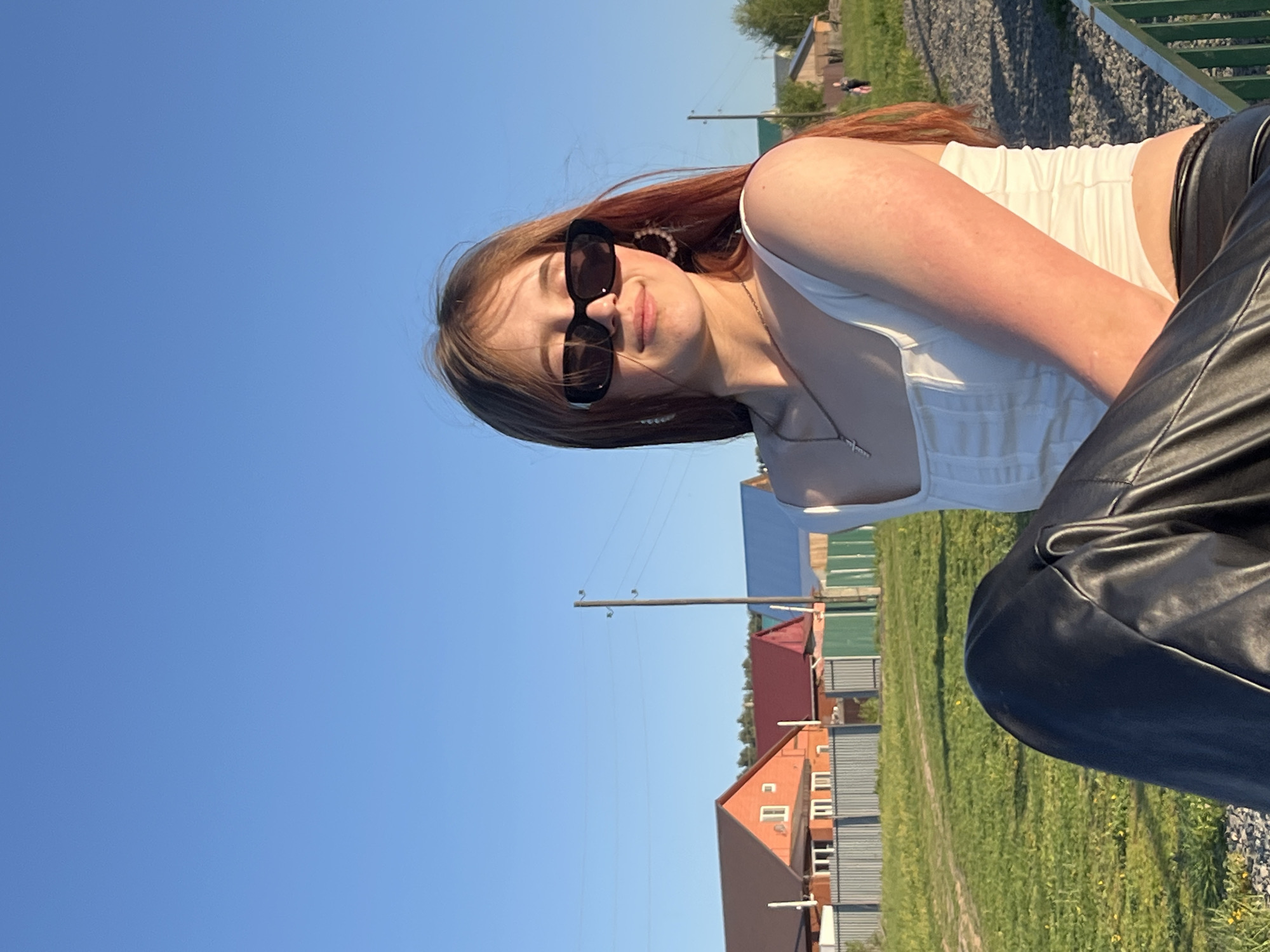 A young woman is posing outdoors in Фоки, Russia. She is wearing a white tank top, black leather pants, sunglasses, and earrings. The background shows houses, grass, and utility poles.