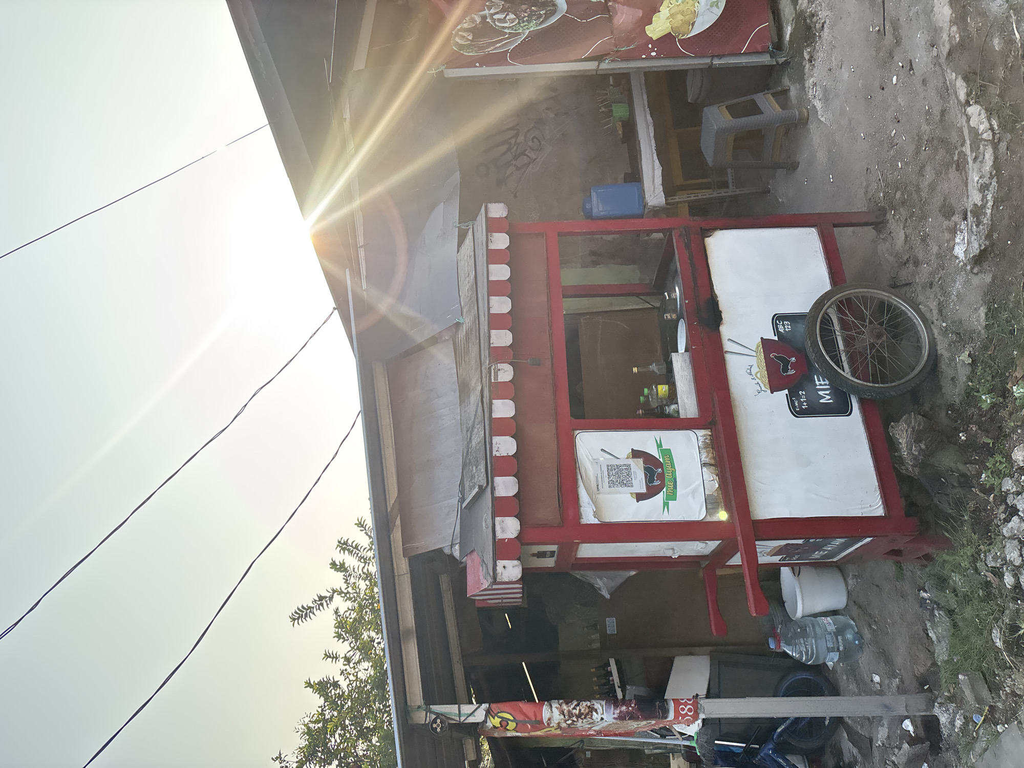 A small, red and white food cart is positioned on a dirt path in front of a modest, open-air structure with a corrugated metal roof. The cart has a canopy with red and white striped fabric. A QR code and a logo featuring a chicken and a bowl of noodles are displayed on the front of the cart, along with the text "mie ayam." A bicycle wheel is attached to the side of the cart. To the left of the cart, a large plastic bottle of water and a white bucket are visible. Behind the cart, a blue plastic water jug sits on a table, along with various bottles and containers. The structure behind the cart appears to be made of rough materials and has graffiti on its wall. A table and a stool are set up behind the food cart. The scene is illuminated by bright sunlight, which casts strong lens flares across the image. Trees with green foliage are visible above the structure on the left side. Power lines are strung across the sky. The overall environment suggests a street-side food stall in a somewhat undeveloped or informal area. This scene is consistent with numerous street food vendors found throughout neighborhoods in West Jakarta, Indonesia, where such carts are a common sight.