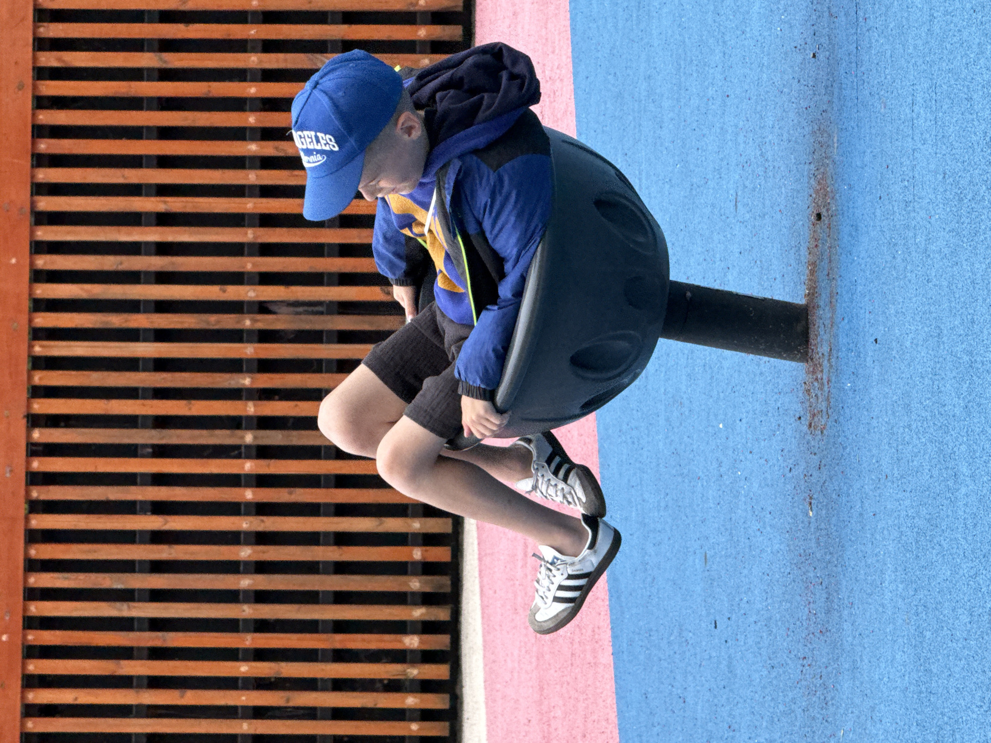 A young boy wearing a blue baseball cap and a blue and yellow jacket is sitting in a spinning playground apparatus. The boy's legs are bent, and his feet are on the ground. He is looking to his right. The playground apparatus is black and has a hole pattern around the rim. The ground is blue and pink. In the background, there is a wooden fence. The image is taken from a slightly low angle. The overall setting suggests an outdoor playground area. The location is not specified in the image, and therefore cannot be determined to be Moscow, Russia.