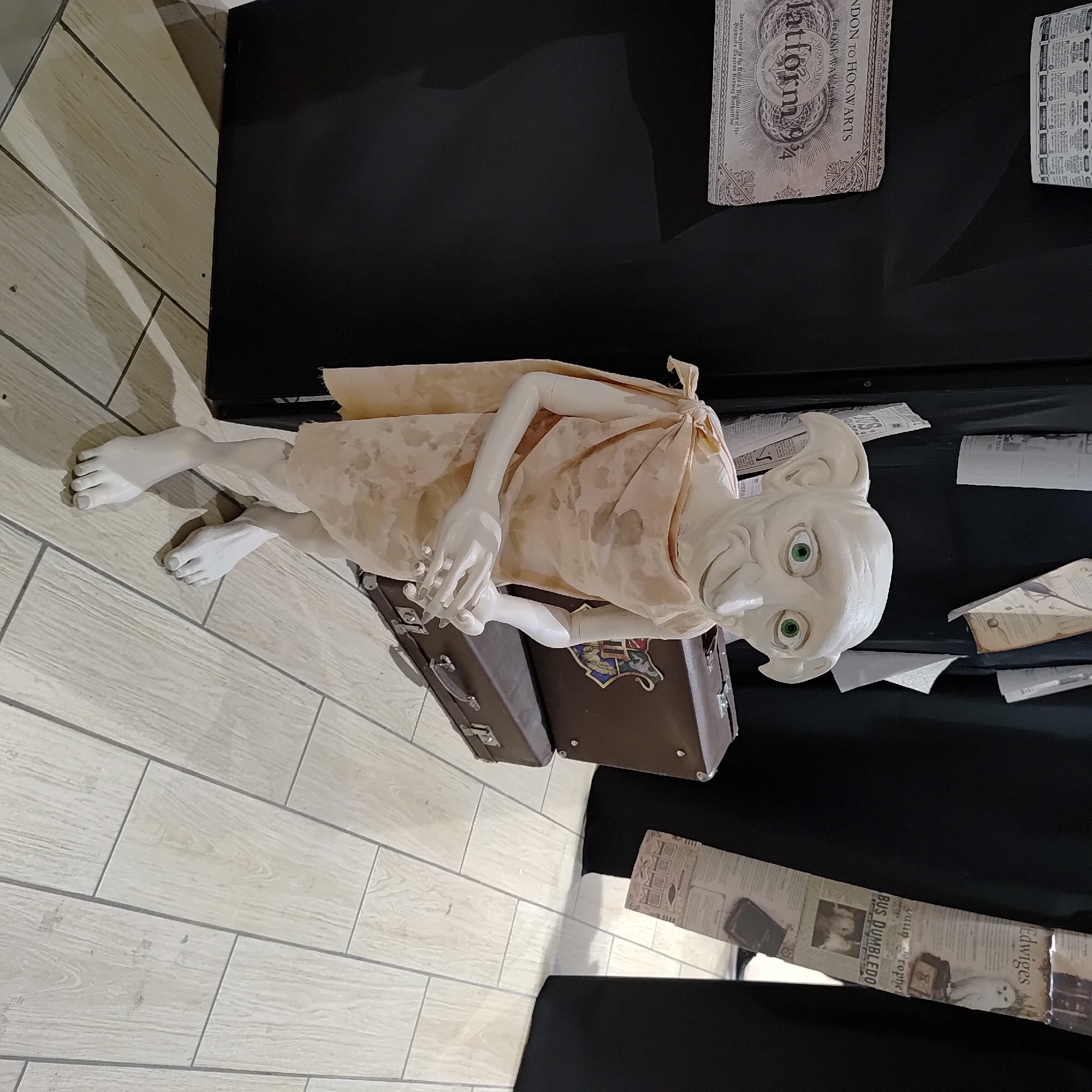 A static, low-angle full shot captures a life-sized model of Dobby, a character from the Harry Potter universe, lying on his side on the floor. Dobby is depicted in his signature large ears, green eyes, and pale skin, draped in a light-colored, patterned cloth. His arms are positioned across his chest, and his legs are bent. Two brown, vintage-style suitcases are placed beneath him, one slightly obscured by his body, and each features a Hogwarts crest.
The floor is tiled with light-colored, wood-effect tiles. In the background, a black surface is visible, with several pieces of paper scattered on it. One piece of paper, placed to the left of Dobby, resembles a Hogwarts Express ticket, featuring the text "Platform 93/4" and "LONDON TO HOGWARTS." Another piece of paper, to the right, is a newspaper-style clipping with text and images, including a picture of an owl with the name "Edwiges" visible. The overall scene is staged, likely in a retail or display setting, possibly related to Harry Potter merchandise. The lighting is artificial, casting shadows that add depth to the scene. No specific time of day or weather conditions are discernible.