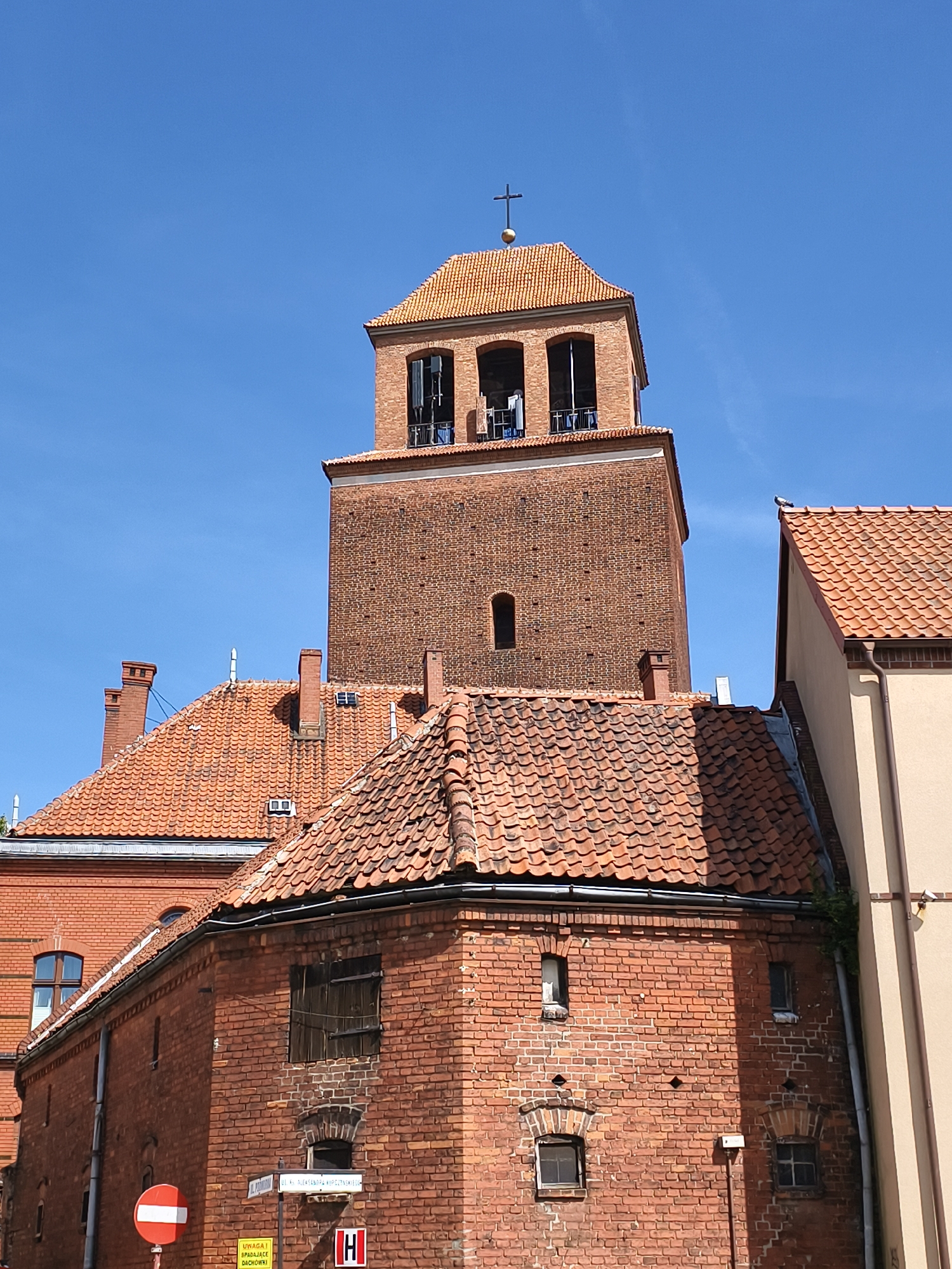 The image shows a close-up view of a historic brick church tower with an orange tiled roof, topped with a cross, against a clear blue sky. The tower is surrounded by other brick buildings with similar tiled roofs. Several windows are visible on the tower and the adjacent buildings, some with wooden shutters. A street sign is partially visible in the lower left corner, and a red and white traffic sign is also present. The architecture suggests it is in a European city. Based on the street sign which reads "UL. ALEKSANDRA FREDRO", this is the Church of Saints Peter and Paul in Tczew, Poland.