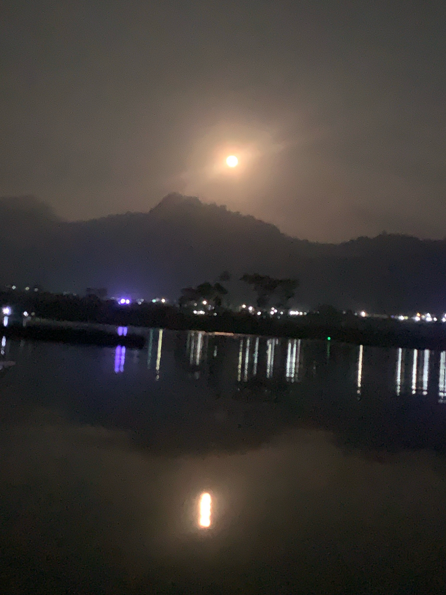 The image depicts a night scene in Kauman, Indonesia, featuring a body of water reflecting lights from the shore and a hazy full moon or sun partially obscured by clouds. The foreground is dominated by the dark water, with blurred reflections of various lights, some appearing as vertical streaks. Along the shoreline in the middle ground, there is a dark silhouette of trees and mountains, with scattered lights suggesting human habitation or activity. The sky is overcast and dim, with a bright, circular light source – likely the moon – creating a diffused glow. The overall impression is one of a tranquil, albeit somewhat indistinct, nocturnal landscape. There are no visible people or specific actions taking place.