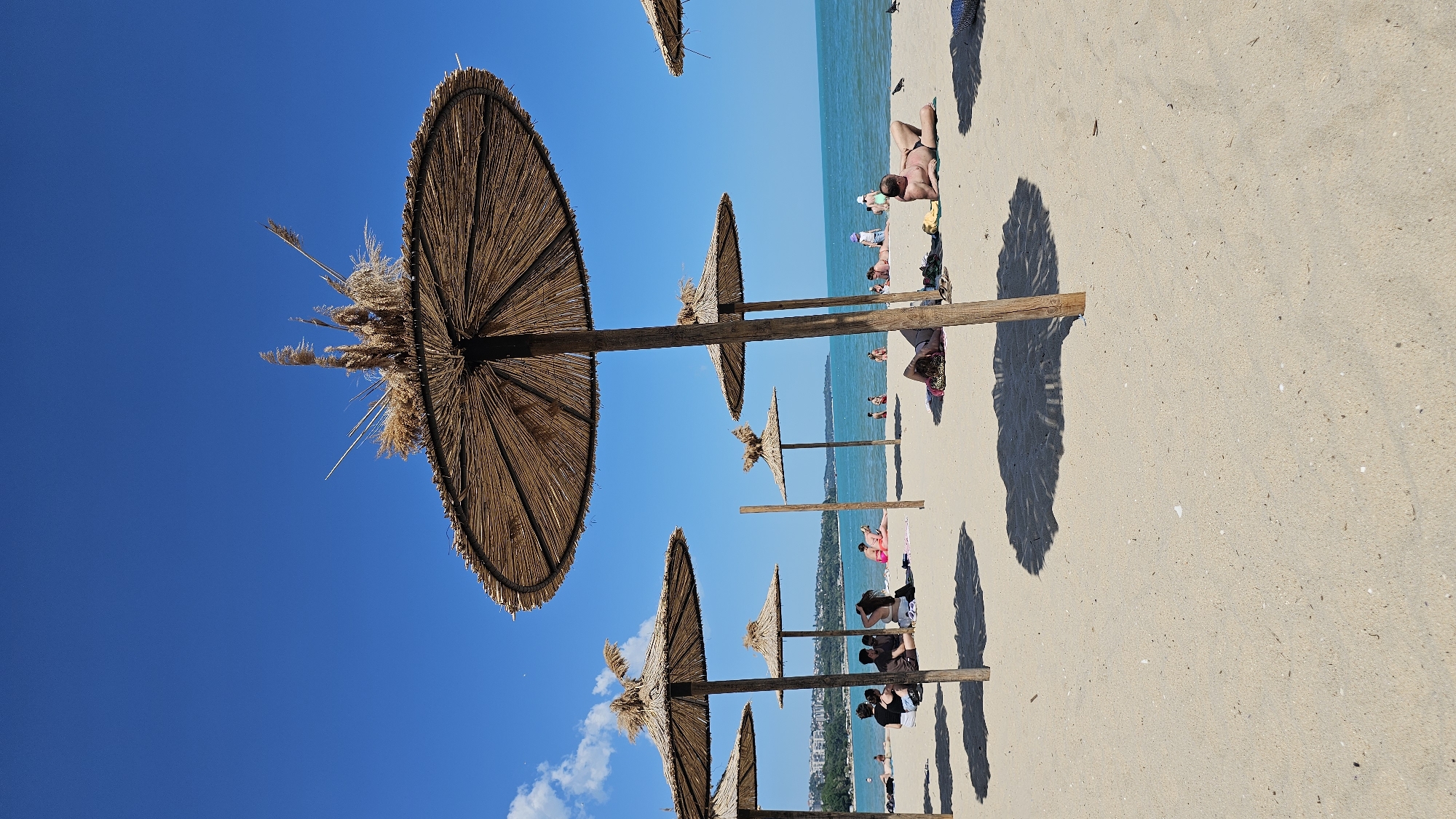 The image depicts a beach scene in Varna, Bulgaria. Several straw umbrellas line the sandy shore, casting shadows on the beach. People are sunbathing and relaxing under the umbrellas. The sea is visible in the background, with a clear blue sky above. The umbrellas are made of straw and have a wooden support structure. The beach appears crowded with people enjoying the sunny weather.