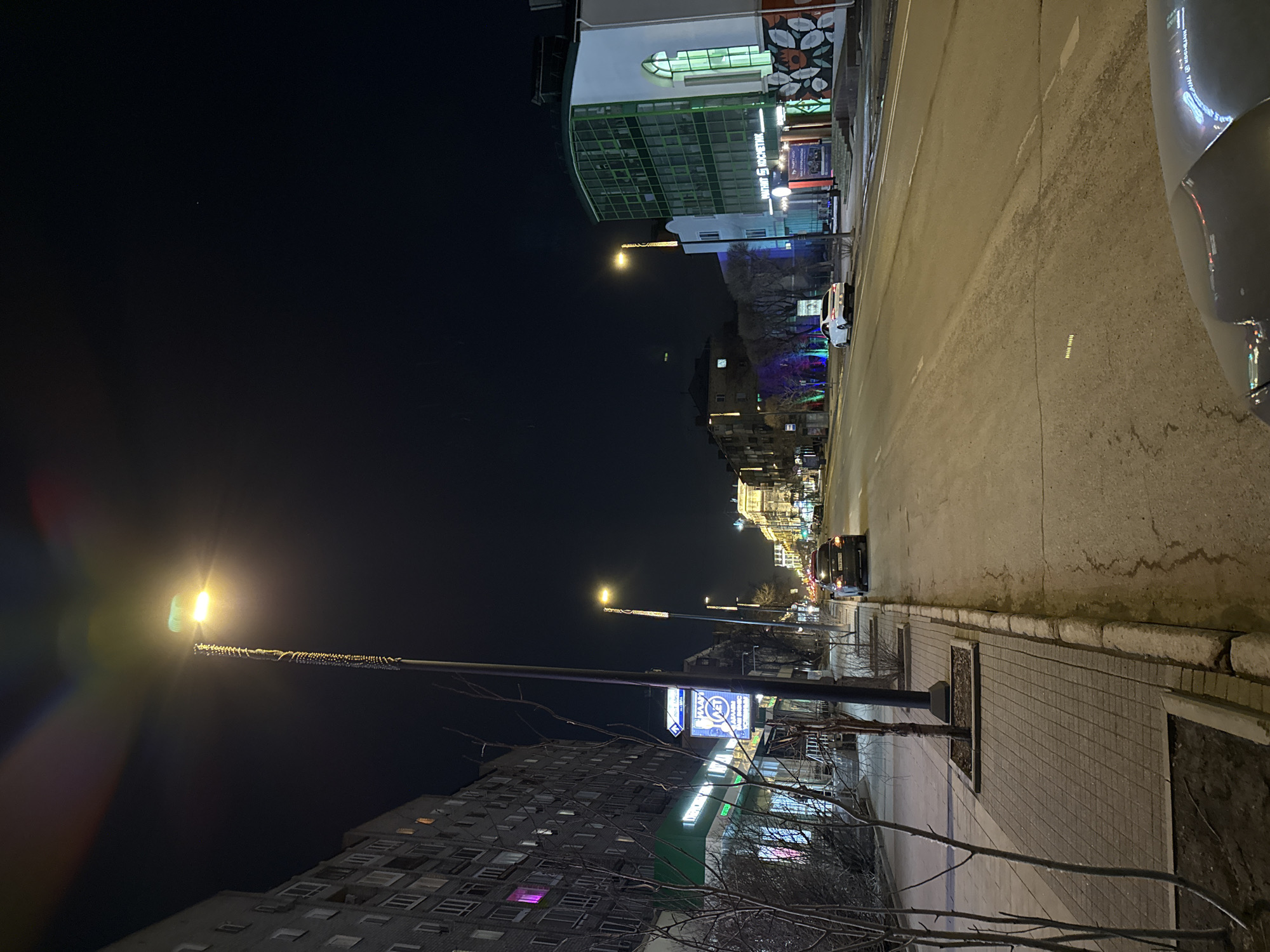 The image presents a nocturnal urban street scene in Krasnoyarsk, Russia, looking down a city thoroughfare lined with buildings. The street is illuminated by numerous artificial light sources, creating a warm, yellowish glow.
On the left side of the frame, a multi-story apartment building with many windows dominates the foreground and midground; one window notably emits a distinct pinkish-purple light. A tall streetlight pole stands prominently in the foreground, adorned with festive lights and emanating a bright, warm light that creates noticeable lens flares. Attached to this pole is a blue and white sign featuring the text "ЛАУНА" (LAUNA) and a stylized 'M' logo with an arrow.
On the right, a series of commercial buildings line the street. A prominent building with a white facade and a green-framed glass section displays the text "ТК ЦЕНТРАЛЬНЫЙ" (Shopping Center Central) and "МАТРОСА ЖЕЛЕЗНЯКА" (Matrosa Zheleznyaka), indicating its name and location on Matrosa Zheleznyaka Street. Another building further down the street is adorned with blue and purple decorative lighting.
The asphalt road, subtly reflective under the streetlights, stretches into the distance, with a few cars visible, their lights glowing. A white car is parked on the right side of the road in the midground, and a dark car is further away. Part of a vehicle is also visible on the extreme right edge of the image. The sidewalk is paved with square blocks, and sparse, bare tree branches and bushes suggest a cold season. The sky above is dark and clear, punctuated by a few faint light specks. The scene conveys a quiet, typical city street atmosphere at night, with no specific activity or people evident.