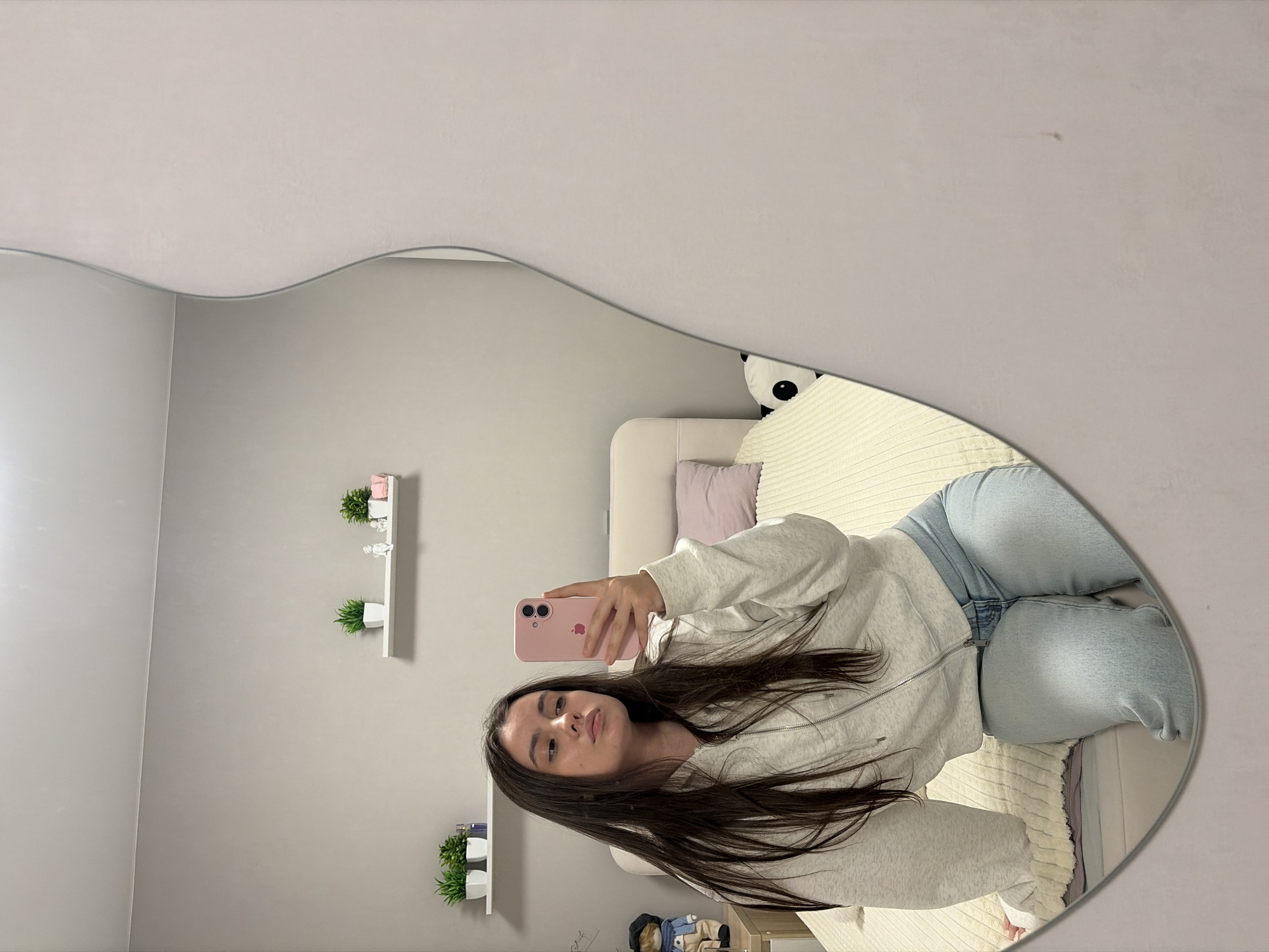 A young woman with long brown hair is taking a selfie in a mirror inside a bedroom. She is wearing a light grey hooded sweatshirt and light blue jeans. She is holding a pink iPhone. The room contains a bed with a light-colored headboard and bedding, decorative wall shelves with small potted plants, and a stuffed panda. Based on available information, the location is Chaikovsky, Russia.