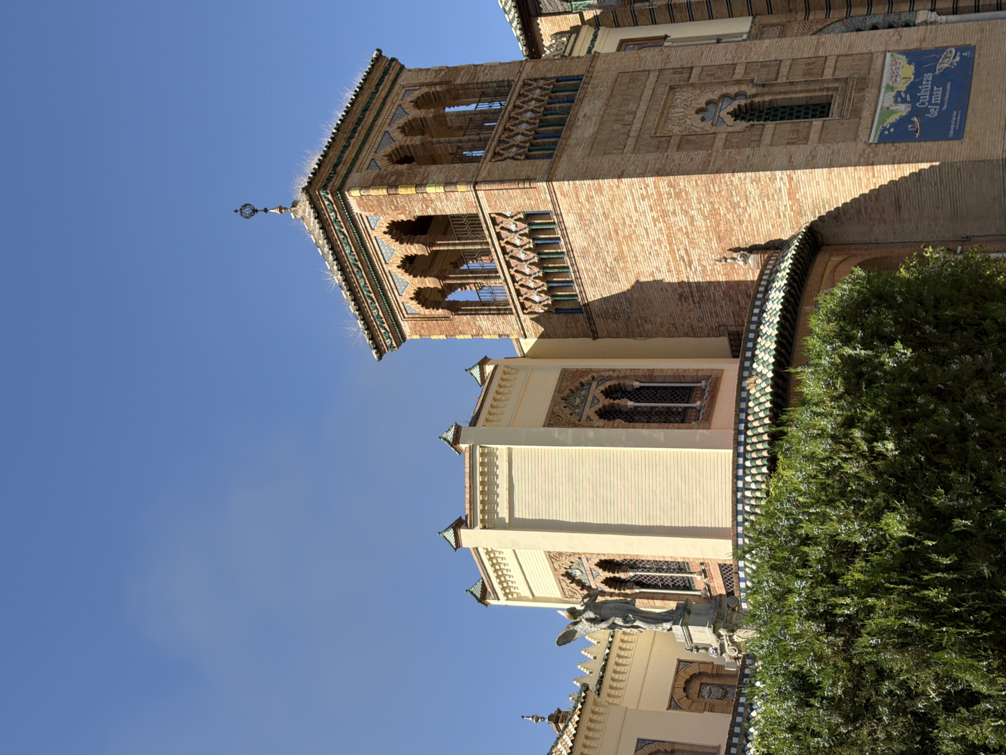 This media file showcases the intricate architecture of a building in Seville, Spain. The building features ornate brickwork, arched windows with decorative metal grilles, and colorful tiled roofs. A prominent sign with the text "Cultura del mar" is visible on one of the walls, suggesting an exhibition or event related to maritime culture. The clear blue sky indicates a sunny day, likely in the morning or afternoon. A lush green tree partially obscures the lower portion of the building, adding a natural element to the scene. The architectural style, with its geometric patterns and detailed ornamentation, points to a Moorish or Mudejar influence, characteristic of Seville. While no people are visible, the presence of decorative elements and the sign imply human activity and cultural engagement.