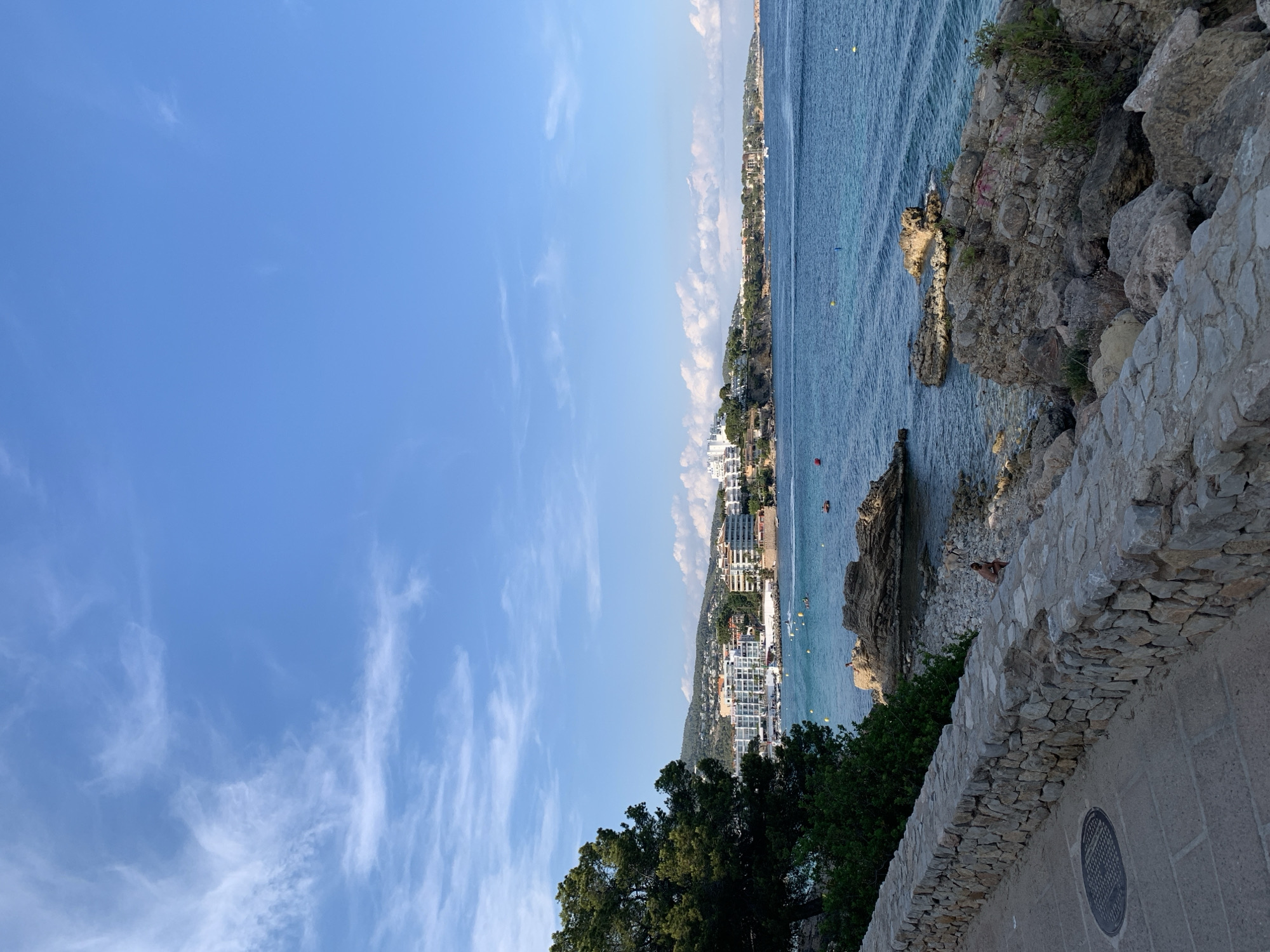 This image captures a scenic coastal view in Palmanova, Spain, under a clear blue sky with scattered wispy clouds. The orientation of the photo is vertical, showing a panoramic scene.
The foreground features a textured stone wall and walkway, with a dark, circular metal grate visible on the pavement. Beyond the wall, lush green trees provide a natural border. The main subject is the vibrant blue Mediterranean Sea, stretching out towards a distant, densely built-up coastline. The shore is characterized by rocky outcrops and a pebbly beach, where a few people are visible, including one person appearing to be sunbathing on the rocks.
Further along the coast, a hillside is covered with buildings, primarily hotels and apartment complexes, showcasing a bustling tourist resort. Several yellow buoys are dotted in the water, suggesting a designated swimming or boating area. A few boats are also visible on the sea, with one leaving a white wake.
The time of day appears to be daytime, likely late morning or afternoon, given the bright sunlight casting shadows and illuminating the scene. The weather is pleasant and clear, ideal for outdoor activities. The overall mood is one of relaxation and vacation, typical of a Mediterranean seaside resort. No visible text is present in the image.