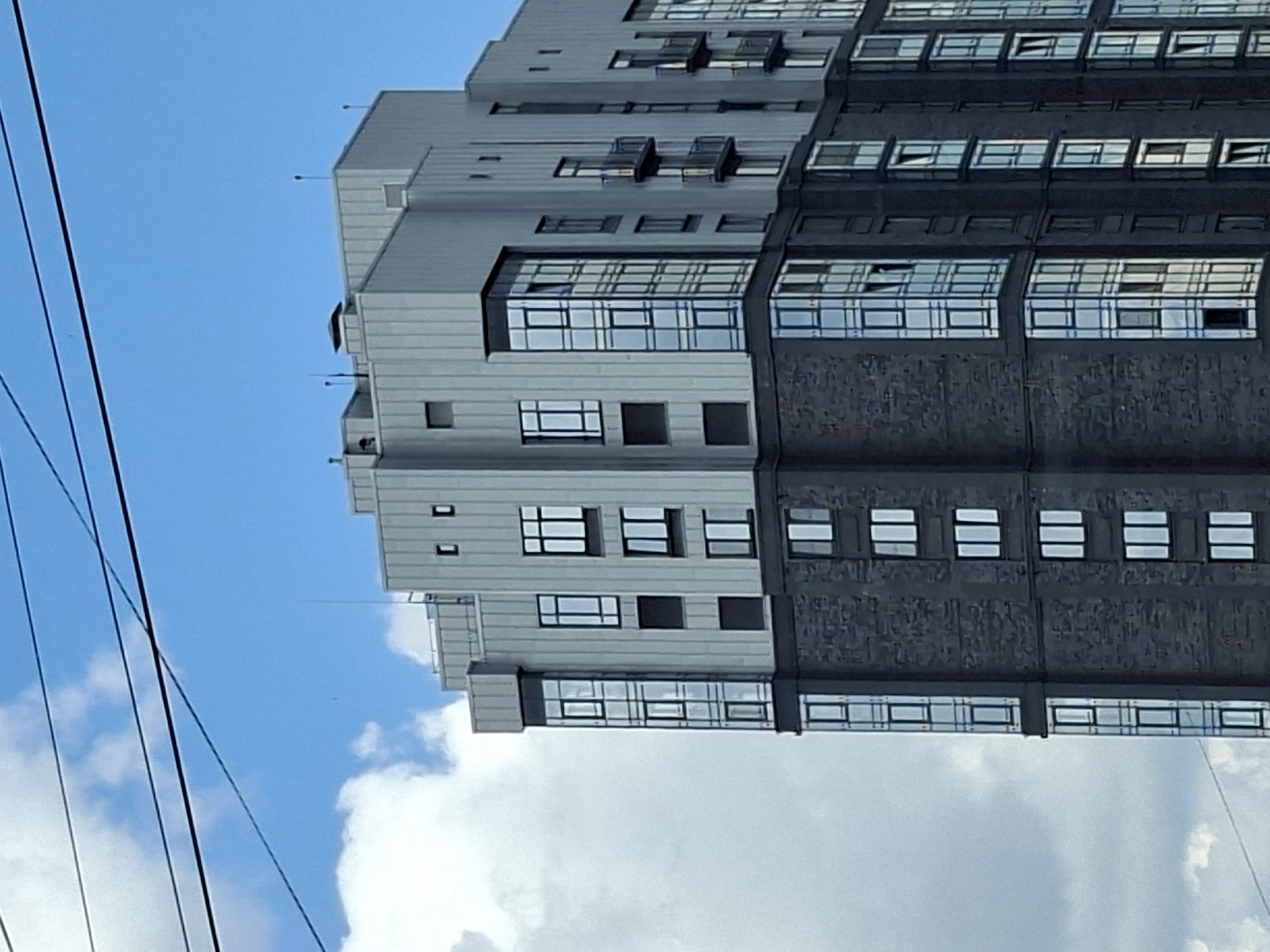 This image, taken in Izhevsk, Russia, shows a modern, multi-story building with a segmented facade. The building is characterized by a mix of light gray panels and dark, textured sections. The windows are predominantly horizontal rectangles, with some appearing to be open. The building's design features several protrusions and recessed areas, creating a varied architectural profile.
The scene is set against a bright blue sky with scattered white clouds, suggesting it is daytime, likely mid-morning or afternoon, given the clear visibility and the illumination of the building. The angle of the photograph, with the building appearing to be viewed from below and to the side, is unusual, possibly due to the orientation of the camera or the placement of the photographer relative to the structure.
Several black wires run diagonally across the left side of the frame, indicating the presence of utility lines in the vicinity. The building itself appears to be residential or commercial, with its uniform window arrangement suggesting individual units or offices. There are no people visible in the frame, and no text is discernable on the building or in the surrounding environment. The overall impression is of an urban setting with contemporary architecture under a pleasant sky.