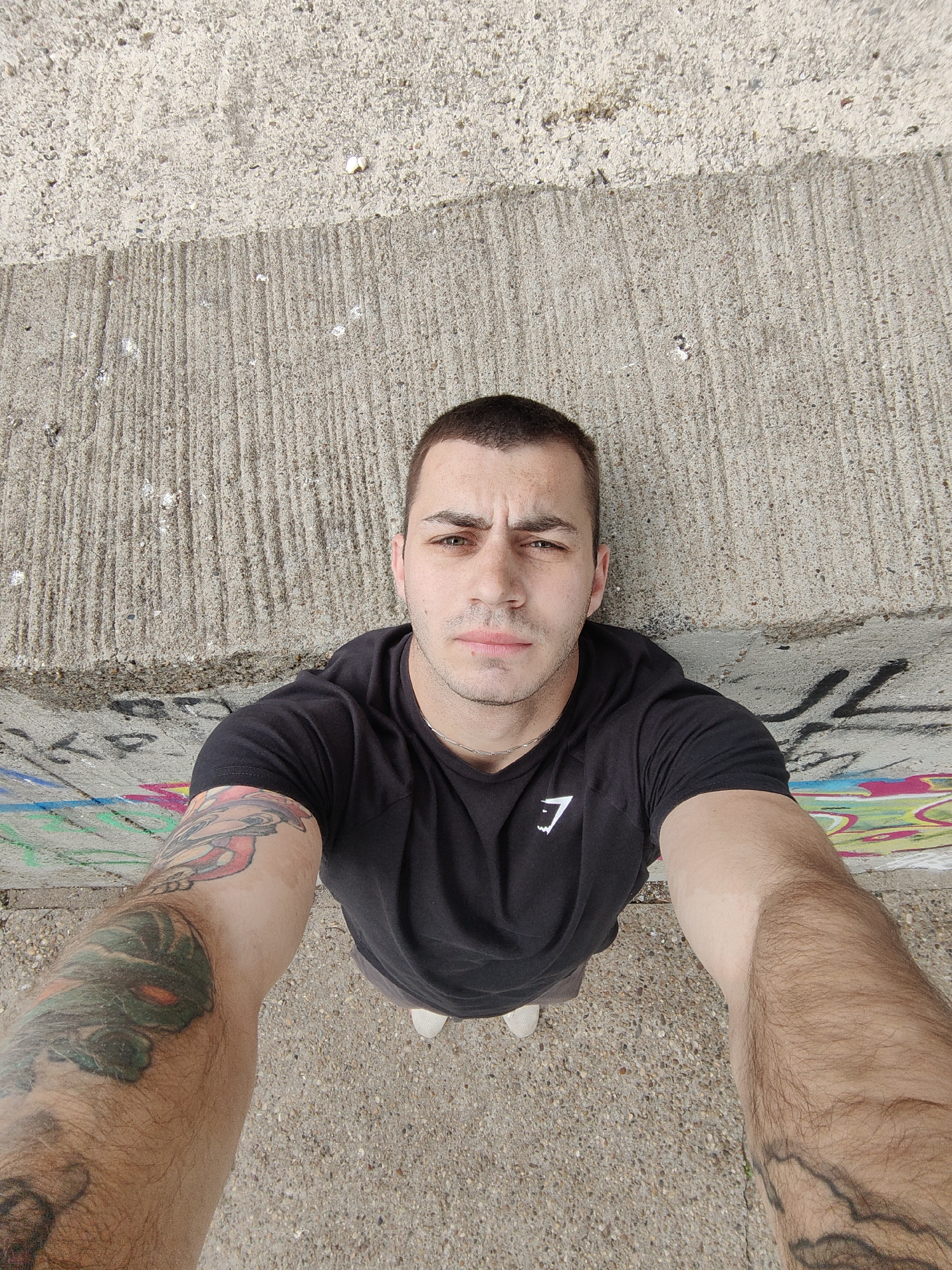 A man is taking a selfie outdoors. He has short, dark hair, light skin, and is wearing a black t-shirt with a white logo. He has tattoos on both arms. The background shows a concrete wall with some graffiti and a textured concrete surface. Based on the graffiti and the overall urban environment, the location is possibly within the city of Varna, Bulgaria, though specific landmarks are absent in the photograph for precise confirmation. The camera angle is from below, looking up at the man.